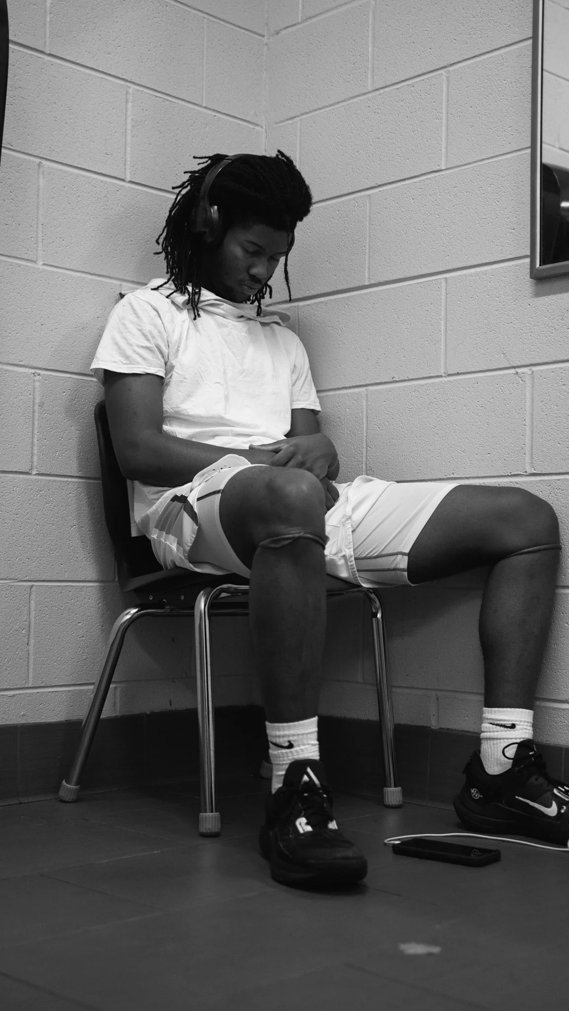 A young man with dreadlocks wearing a white shirt and shorts, sitting on a chair in a room with cinder block walls, listening to music with headphones, with his shoes placed on the floor in front of him.