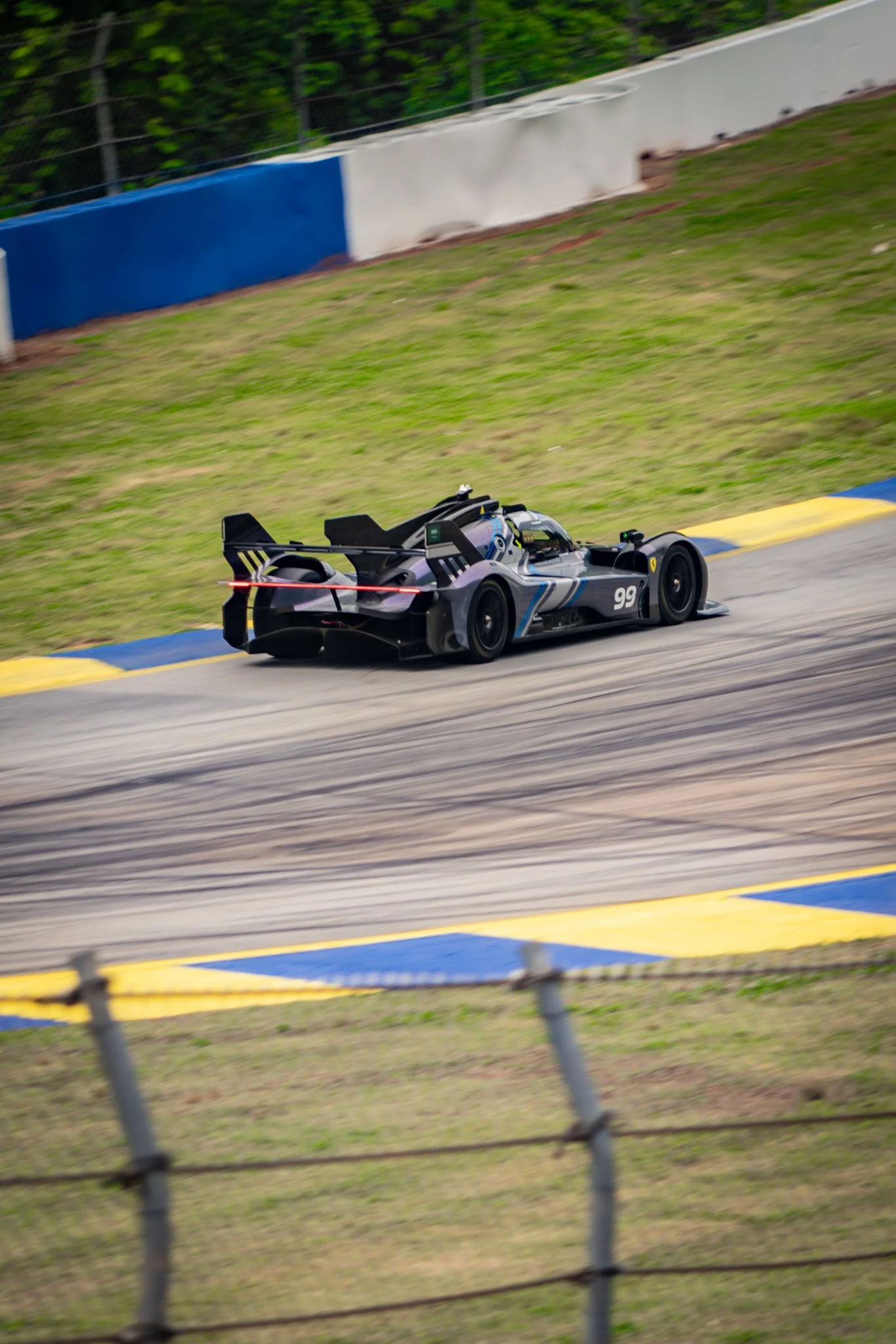A black race car with blue and white accents driving on a race track with a grassy area and barriers on the side.