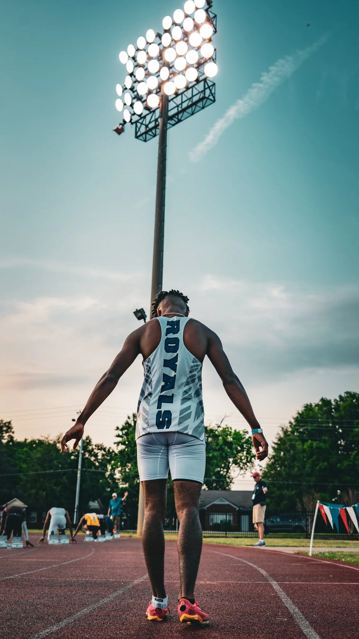 Track athlete in a relay race preparing to run under stadium lights, with other runners and officials in the background.