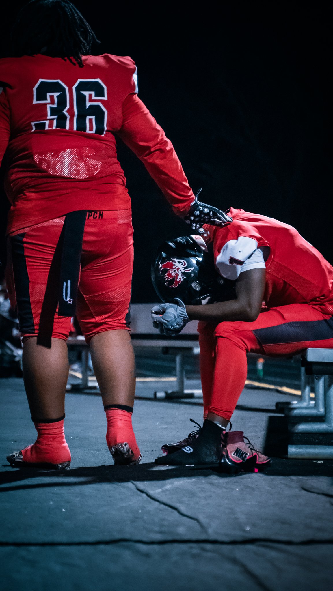 Two football players wearing red uniforms, one is sitting on a bench with a helmet and gloves, the other is standing with a jersey number 36, at night under artificial lighting.