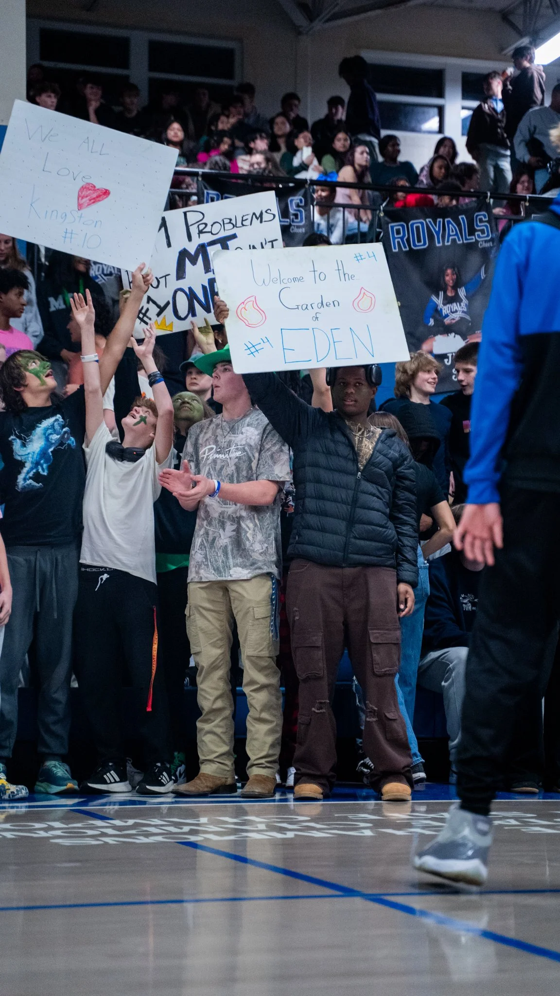 A group of students at a school assembly holding various handmade signs, including one that reads 'Welcome to the Garden of EDEN' and others with messages of love and school pride, in a gymnasium with spectators and banners in the background.