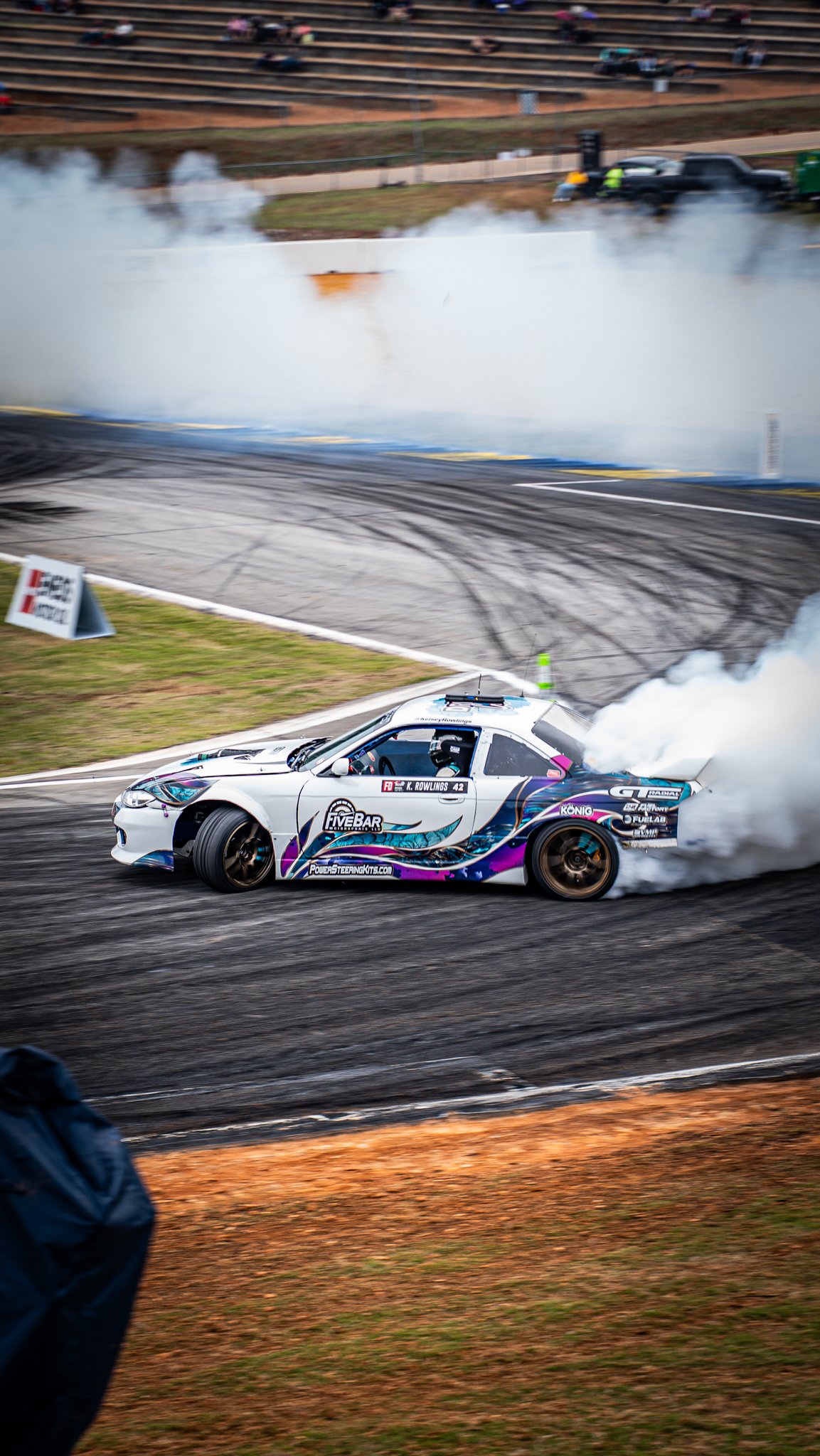 A white racing car drifting around a corner on a race track, generating smoke from its tires, with tire marks visible on the asphalt. Background includes empty bleachers and a person watching.