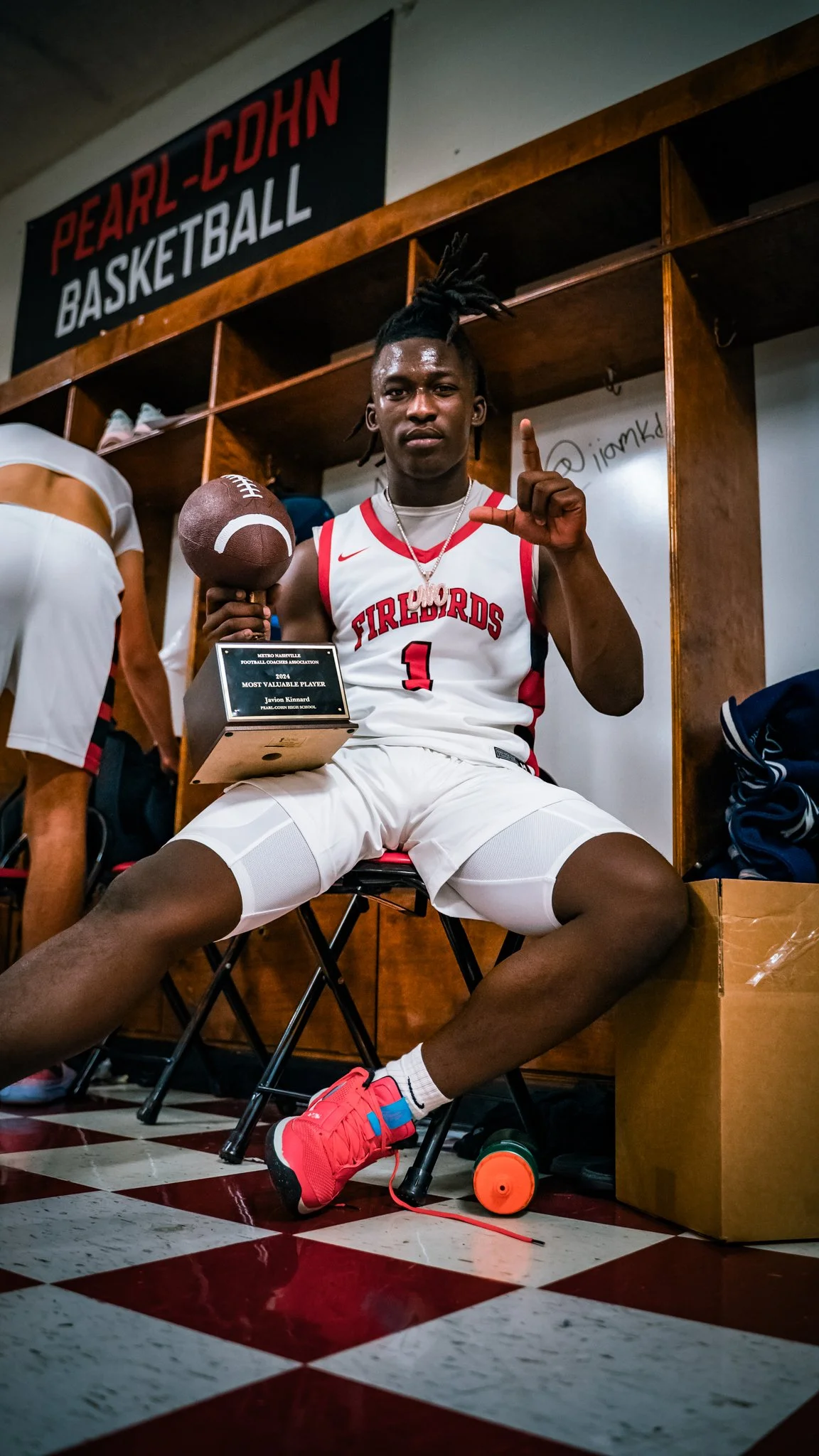 Young male basketball player sitting in locker room, holding a football, wearing a white basketball uniform with red accents, making an 'L' shape with his hand, and showing a plaque for most valuable player award.