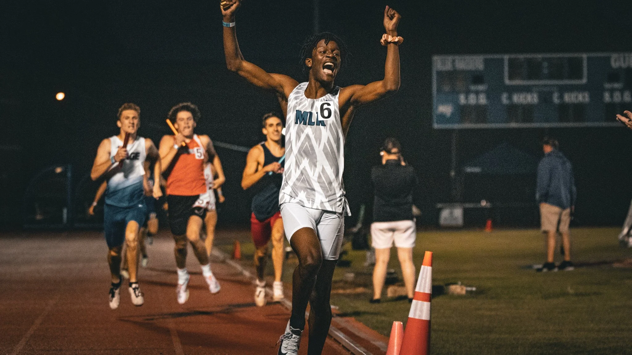 A group of runners competing in a race at night, with the lead runner raising his arms in victory near orange cones on a track.