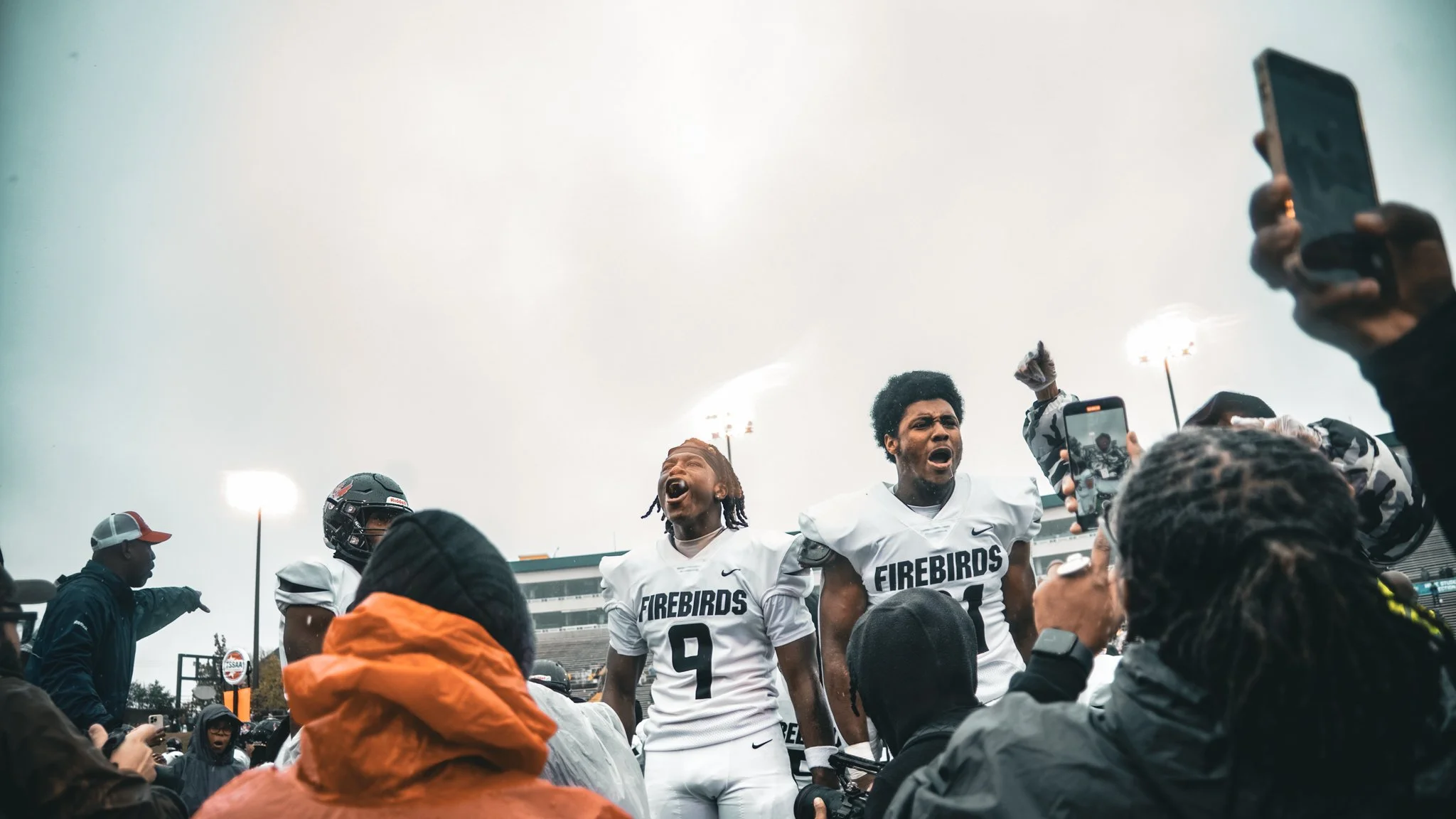 A group of football players in white jerseys with 'Firebirds' written on them celebrating on the field, surrounded by fans and photographers during a game under cloudy skies.
