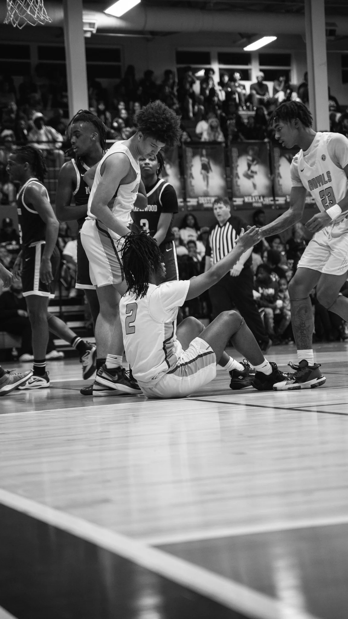 Black and white photo of a basketball game. A player wearing jersey number 2 from one team is sitting on the floor reaching out to another player wearing jersey number 23 from the opposing team, who is extending his hand. Several other players and a 