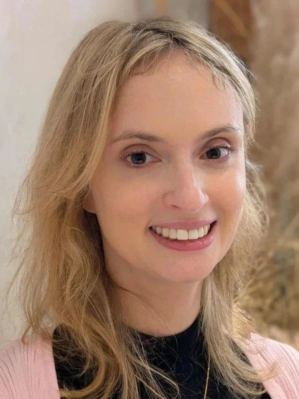 A young woman with shoulder-length blonde hair, blue eyes, and fair skin smiling at the camera. She is wearing a black top and a pink cardigan and is indoors with a wooden and beige background.