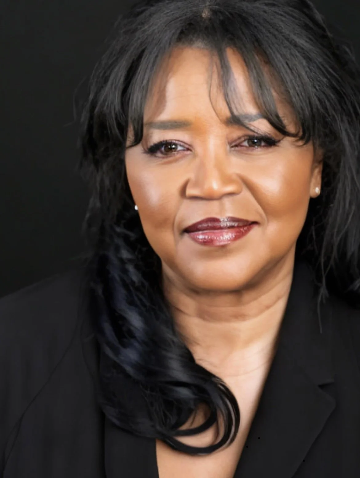 A portrait of a middle-aged woman with dark, wavy hair, wearing a black blazer, with a subtle smile and a dark background.
