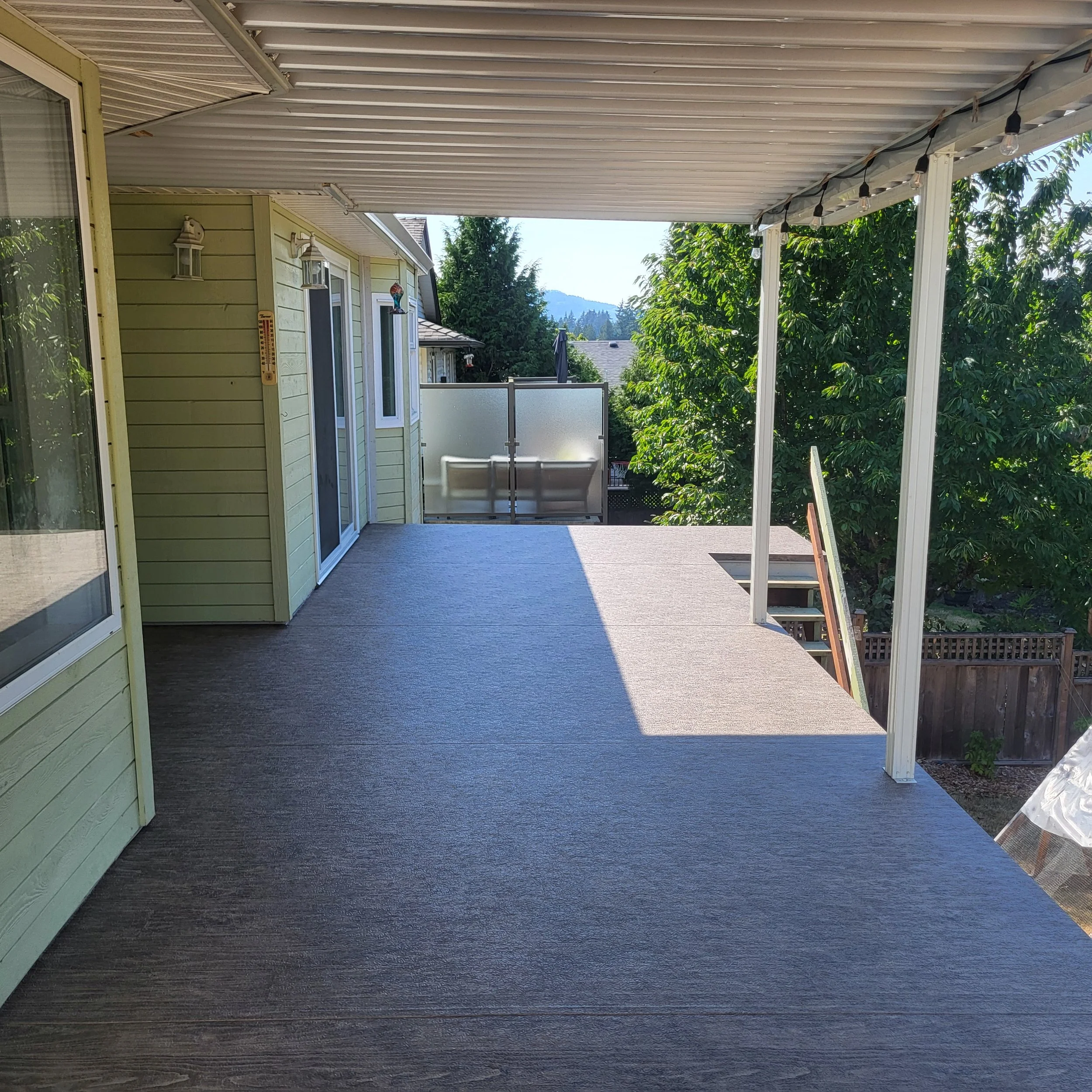 View of a balcony or porch area with a textured gray floor, beige siding, glass doors, decorative lanterns, and string lights. A mountain and trees are visible in the background.