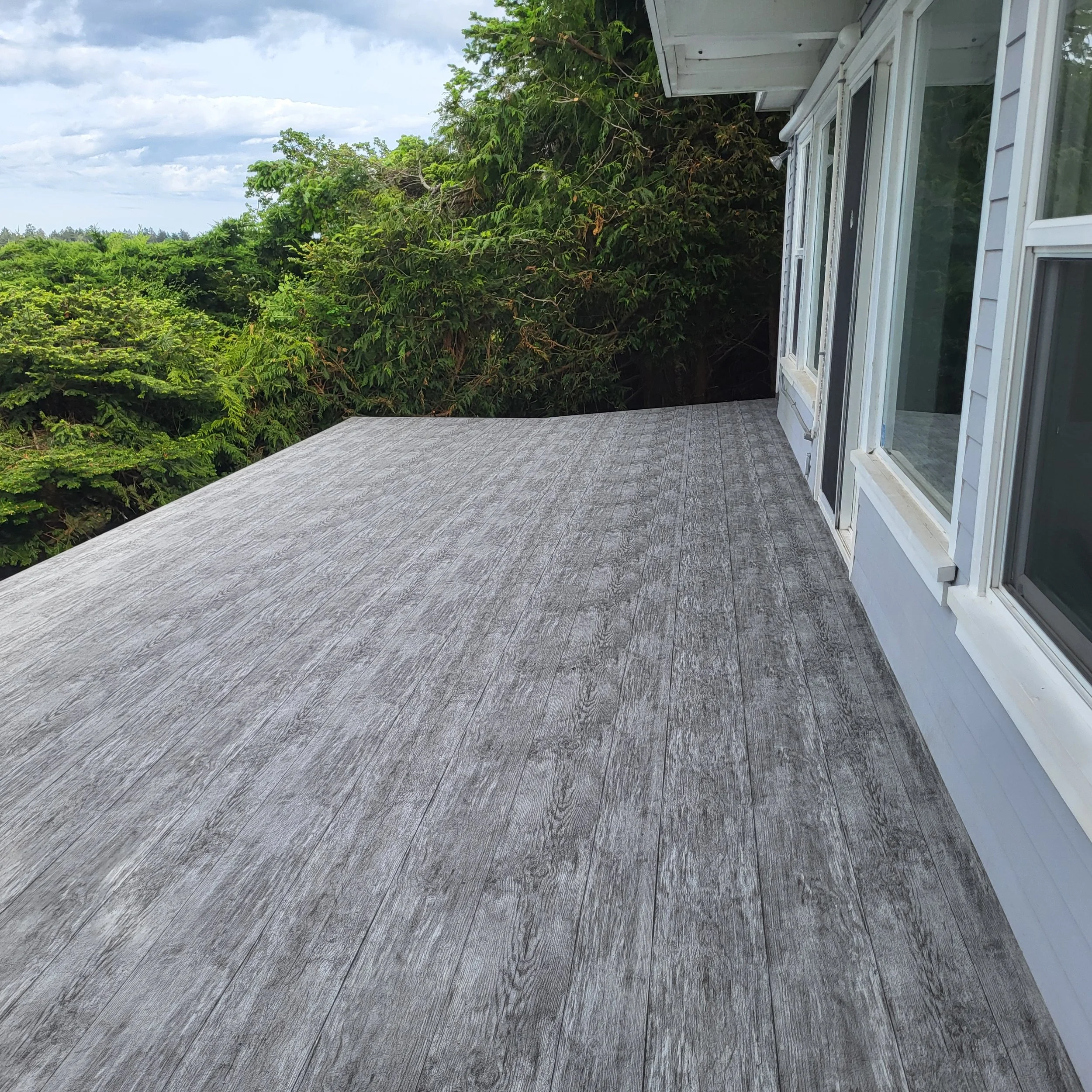 A wooden deck extends from a house with white siding and large windows, overlooking green trees and a cloudy sky.