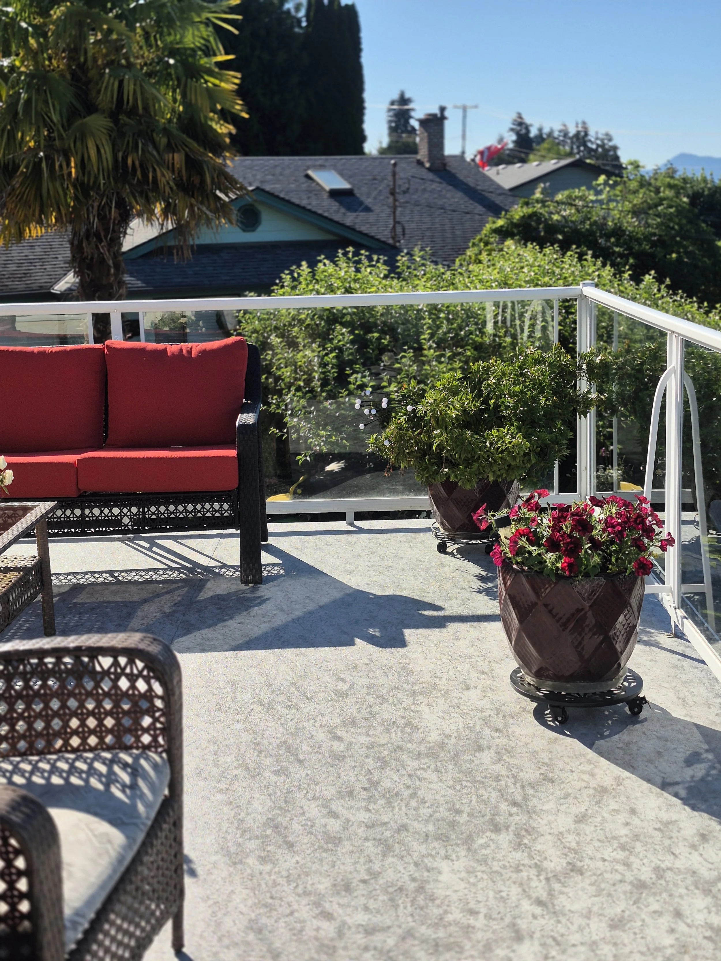 A porch with chairs, potted plants, and a glass railing, overlooking houses with trees and mountains in the background on a sunny day.