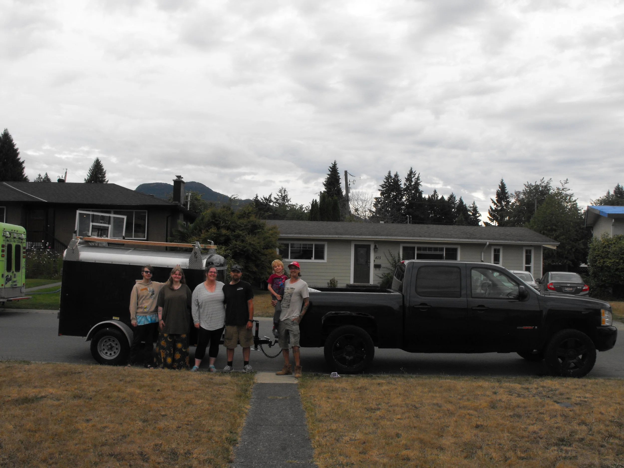A group of seven people, including children and adults, standing in front of a black trailer on a residential street on a cloudy day. There are houses, trees, and mountains in the background.