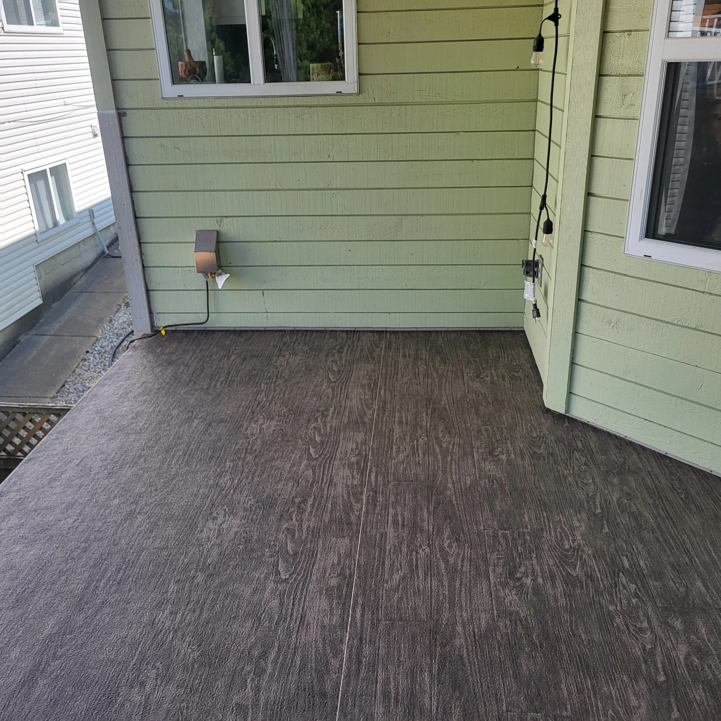 Backyard patio with green wooden siding, double glass doors, outdoor lighting, and electrical cords on the deck, with neighboring house visible in the background.