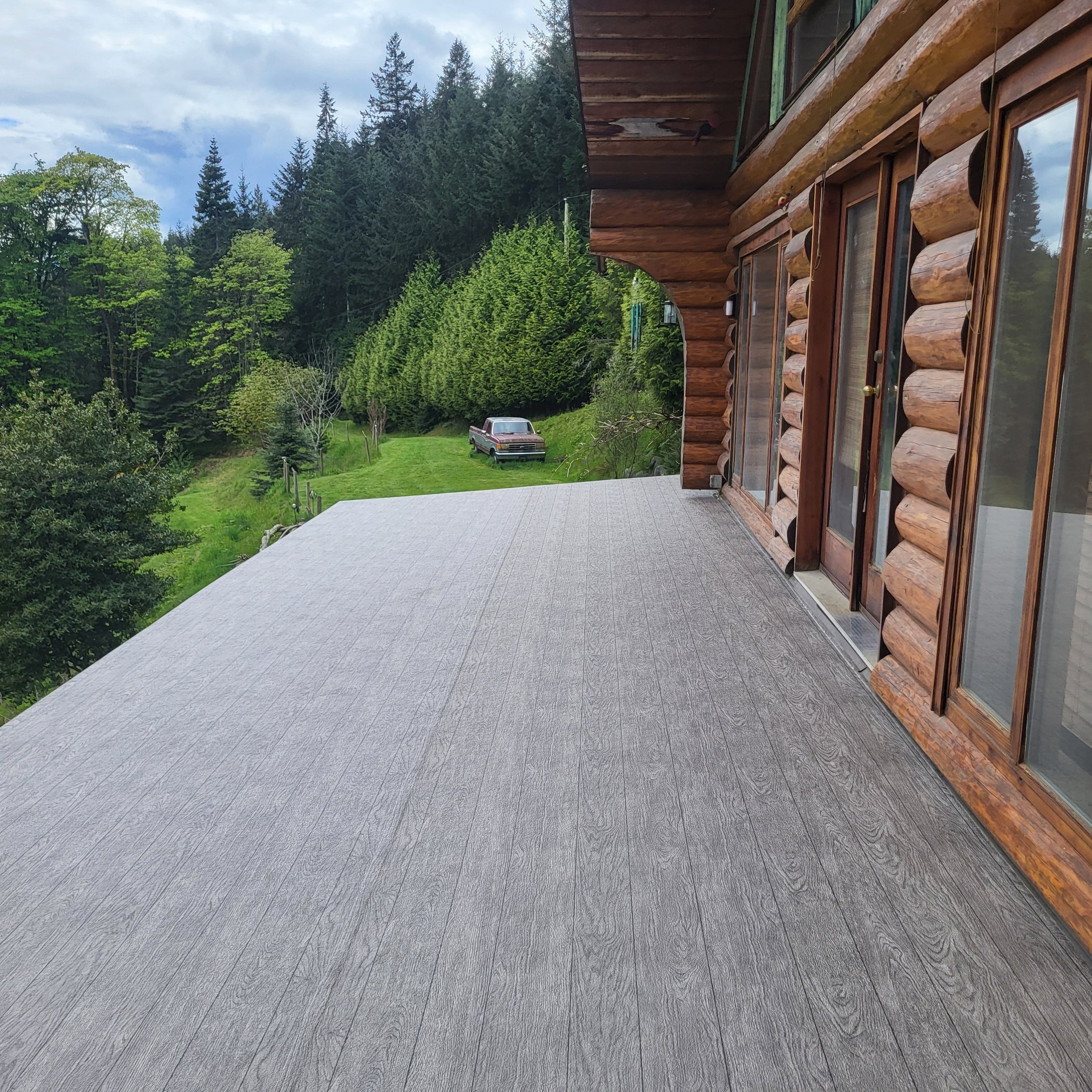 Completed grey vinyl deck beside a log home, with a clean waterproof surface overlooking a wooded yard.
