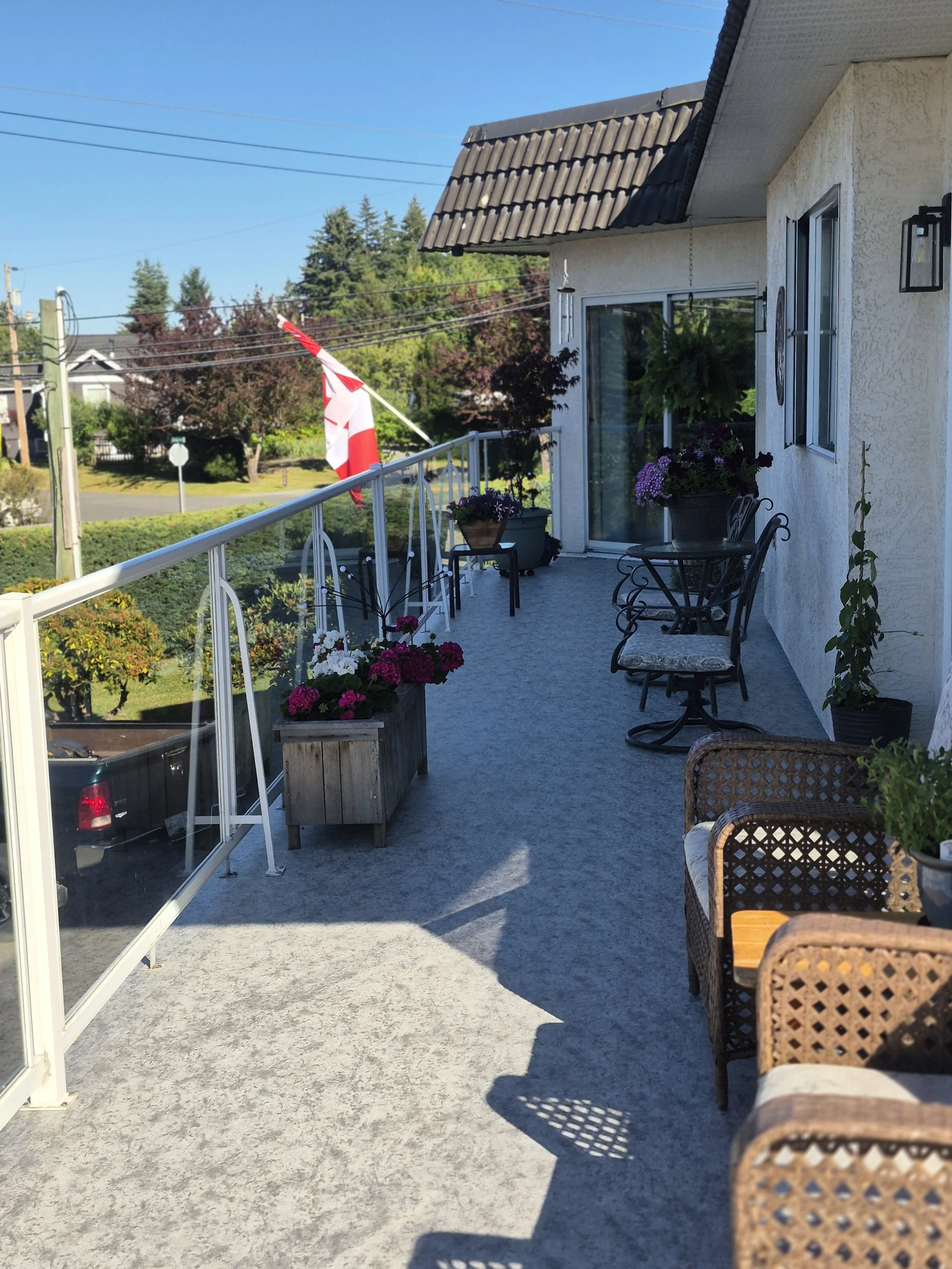 A balcony with patio furniture, potted flowers, and a Canadian flag, overlooking a suburban neighborhood with trees and houses under a clear blue sky.