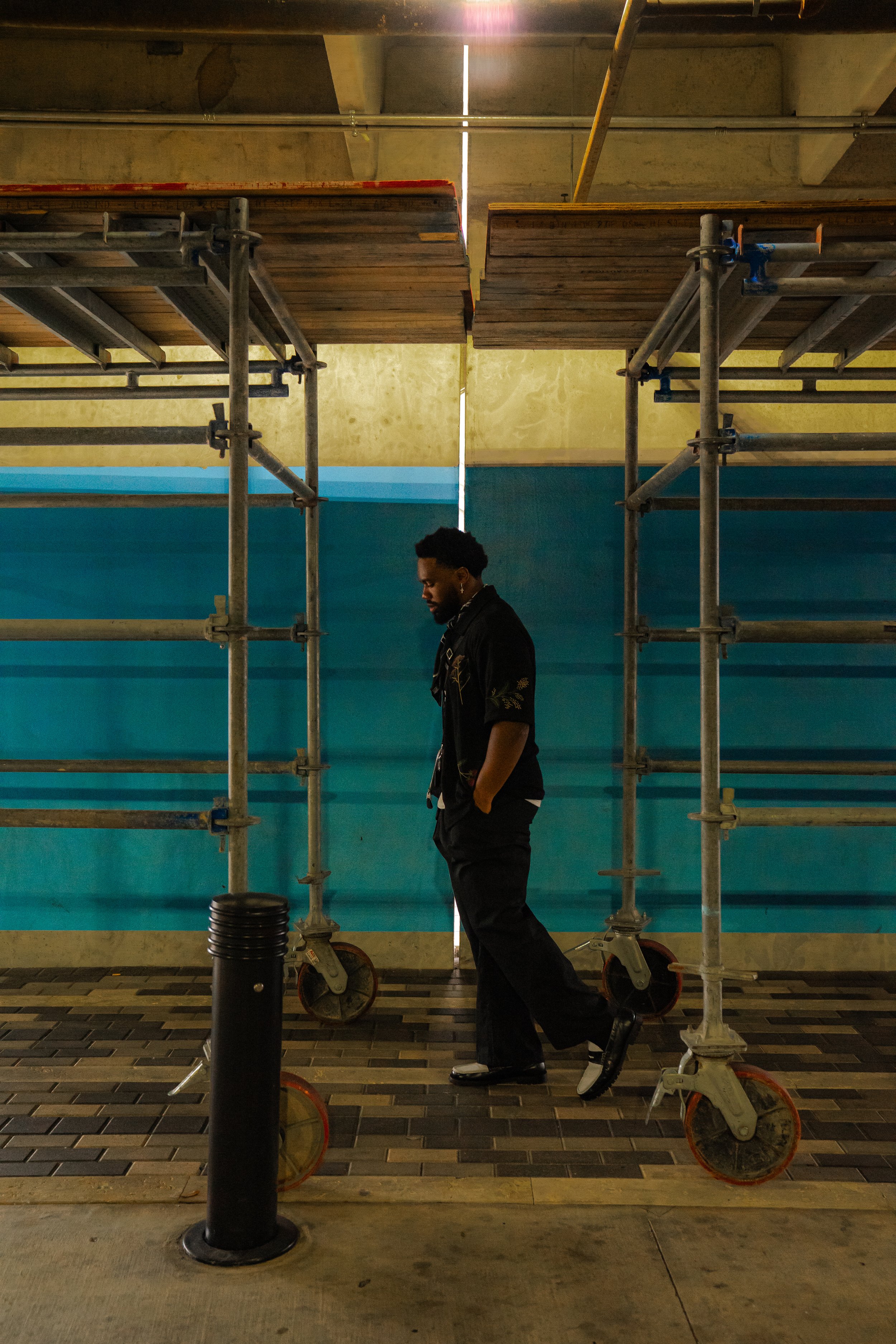 “Jacob Berry posing in a dark streetwear look beneath an industrial structure with blue paneling.”