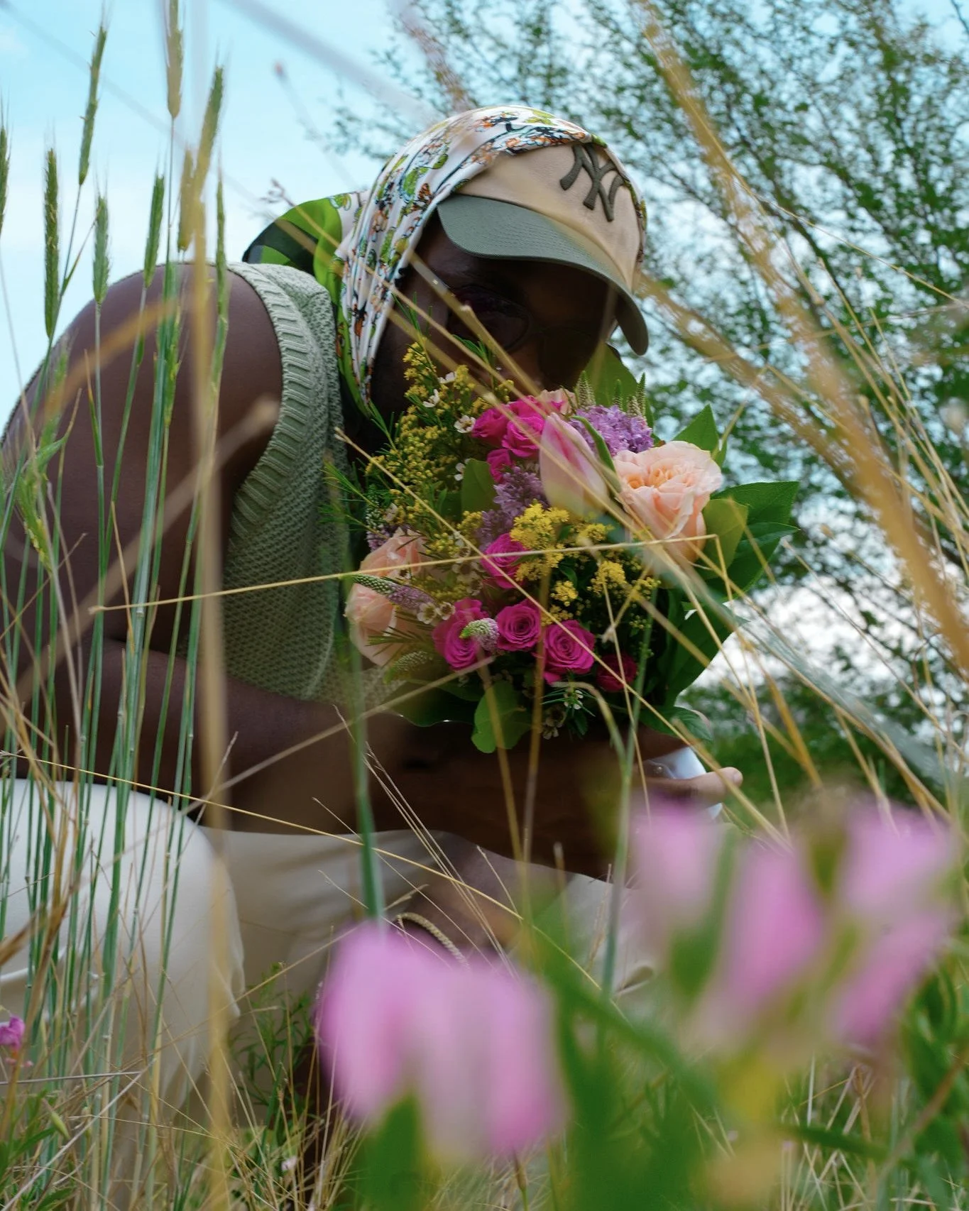 Flowers: “Jacob Berry holding a colorful bouquet while wearing a knit vest, baseball cap, sunglasses, and patterned headscarf in a grassy field.”