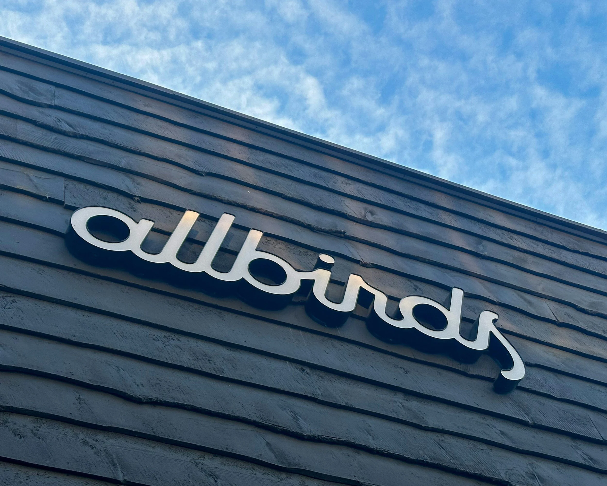 Close-up of a building's wall with horizontal gray siding and a partial view of the word 'Chelsea' in silver letters, under a blue sky with scattered clouds.