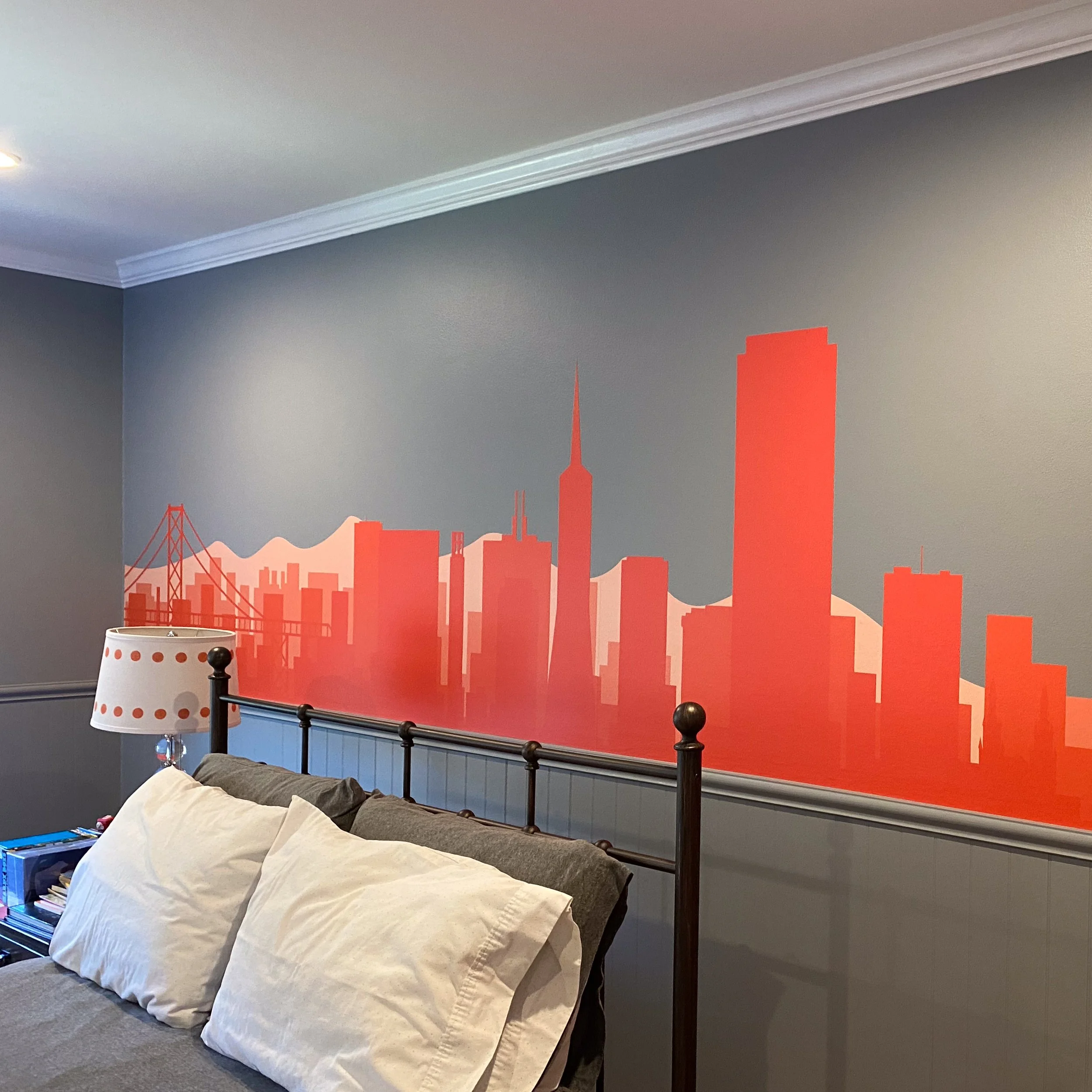 A bedroom with a black metal bed frame, a beige blanket, and a pillow on the bed. A lamp with a white shade and orange dots sits on a nightstand, and there are books and other items on it. The wall behind the bed features a bright orange city skyline mural, including tall buildings and a bridge, set against a bluish-gray wall.