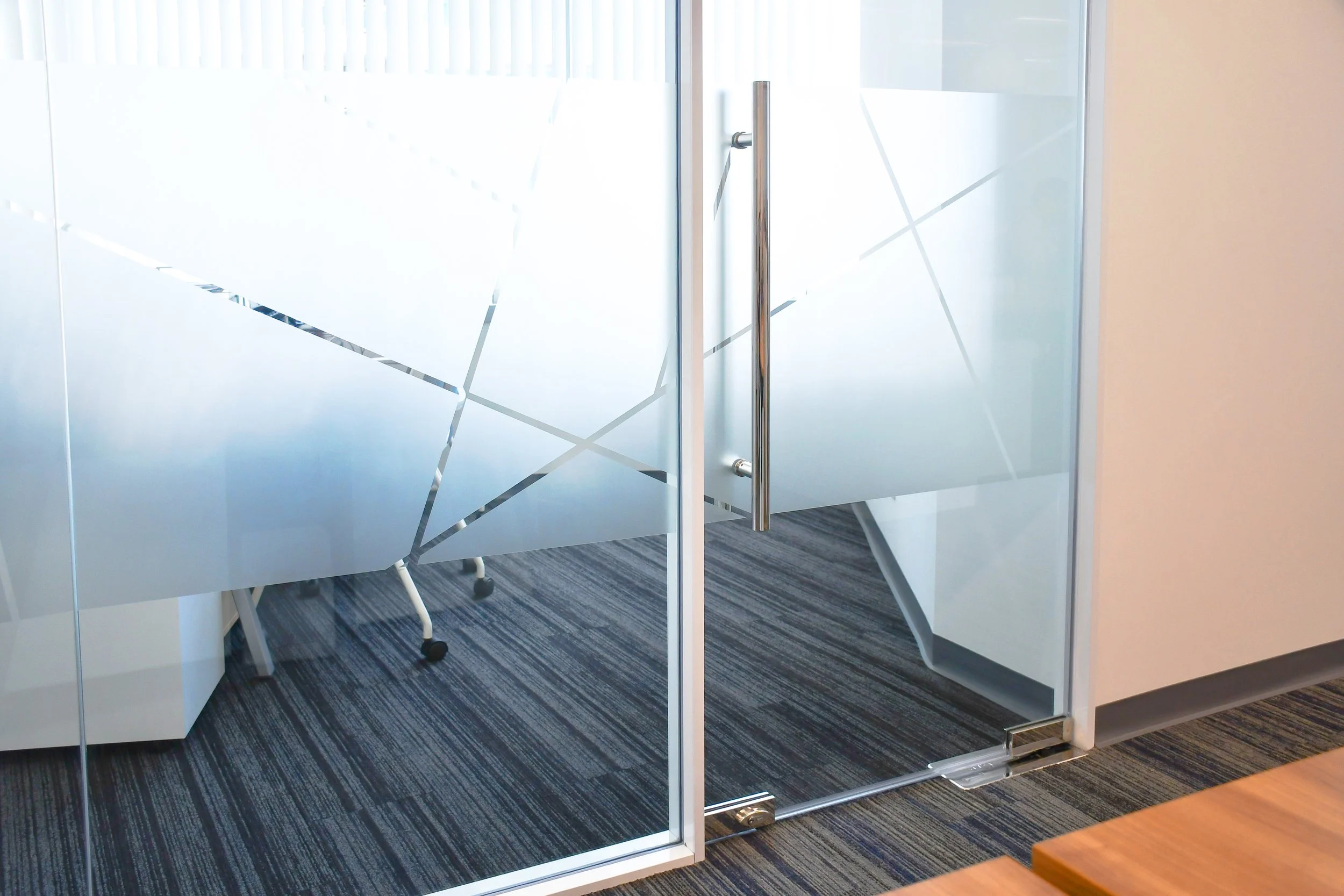 Frosted glass office divider with geometric patterns and a glass door with a metal handle, in a modern office setting with a carpeted floor.