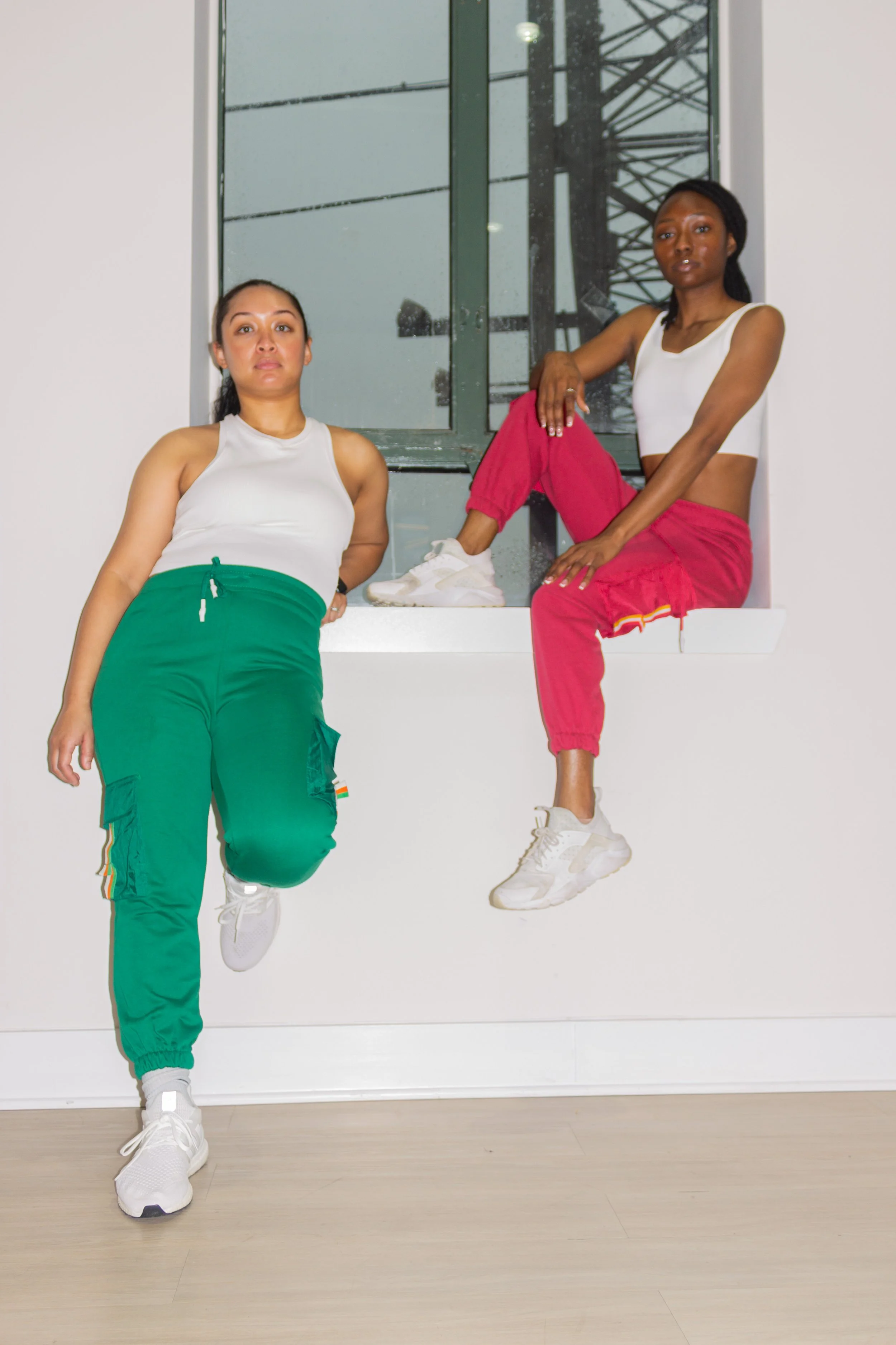 Two women with dark hair in white tops and colorful joggers, wearing white sneakers, posing in front of a large window with a tower structure visible outside.