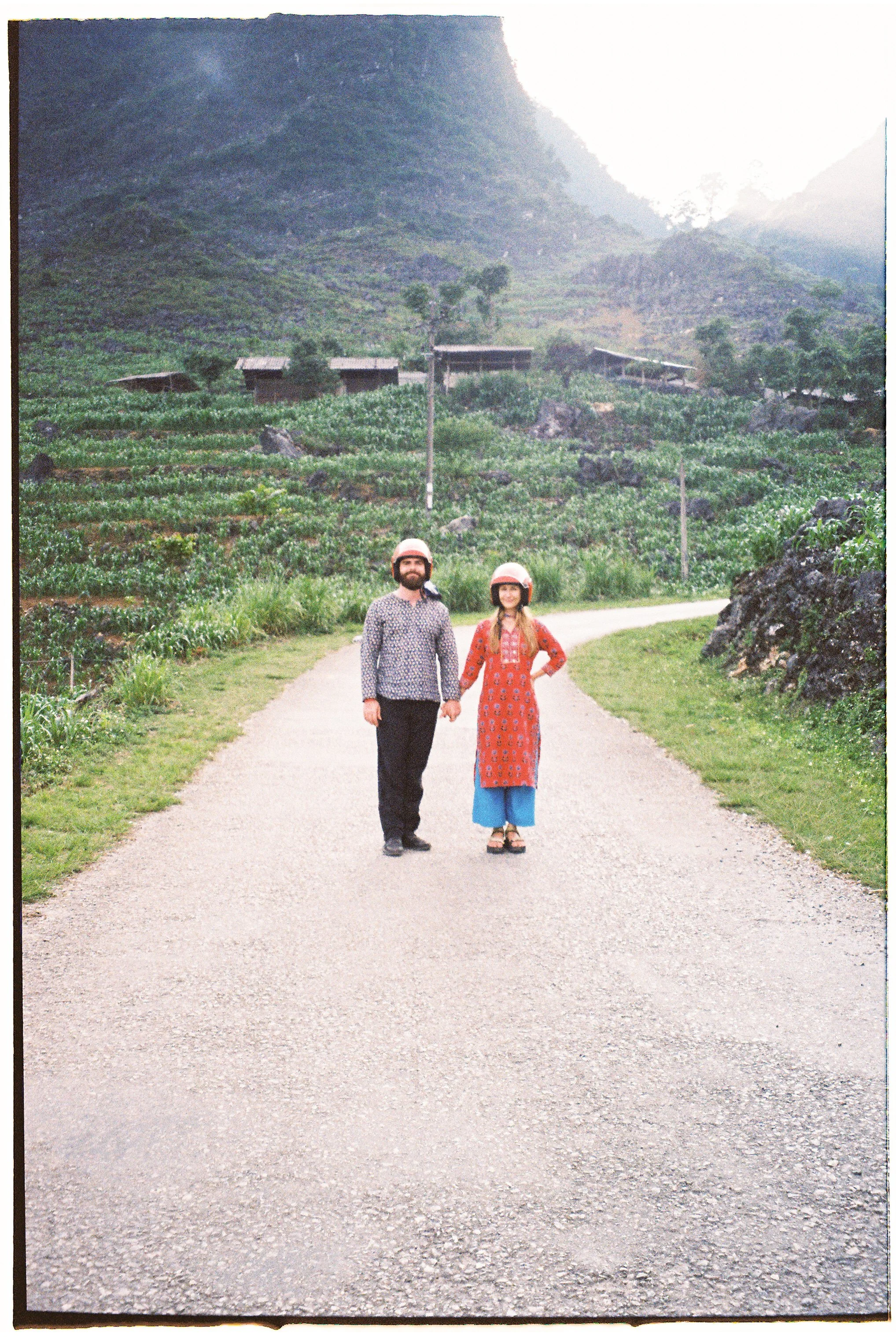 A man and woman wearing helmets and holding hands walking on a gravel road in a rural area with green fields, small houses, trees, and a mountain in the background. A film Portrait of Eli and Olivia Ladesic in Vietnam.