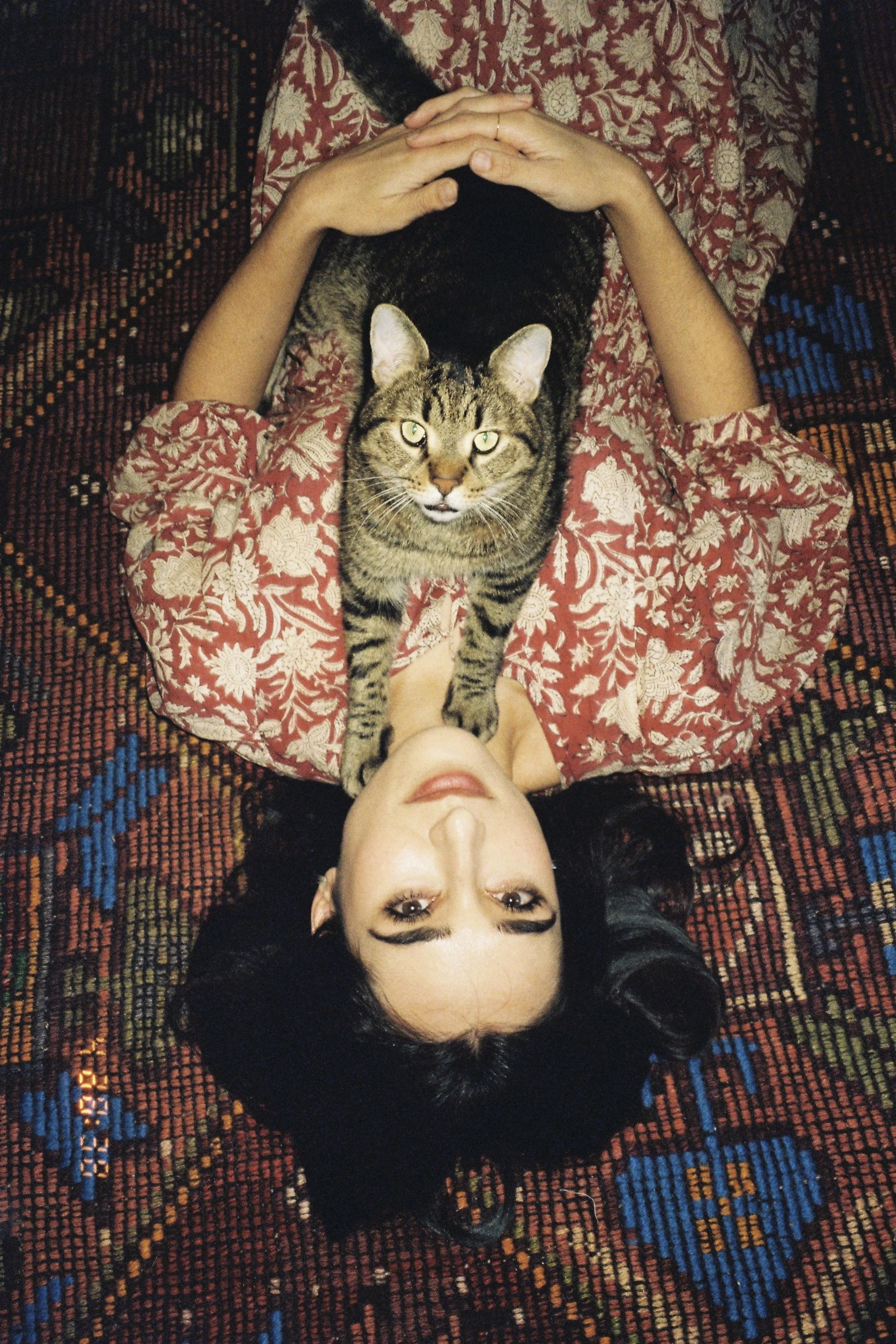 A woman with dark hair and makeup lying on her back on a patterned rug, with a tabby cat stretched across her chest, looking up at the camera. Film shot taking at a branding shoot in Tbilisi, Georgia.