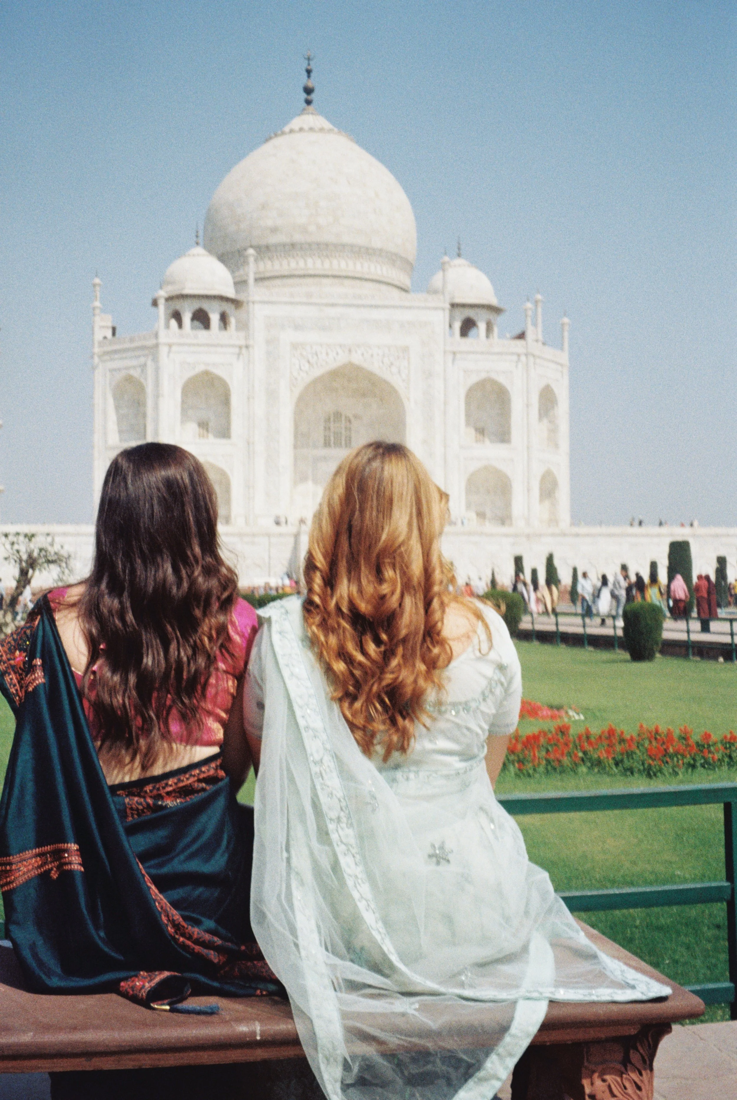 Two women with long hair, one dark and one light, sitting outside in front of the Taj Mahal. Film shot by Olivia in Agar, India.