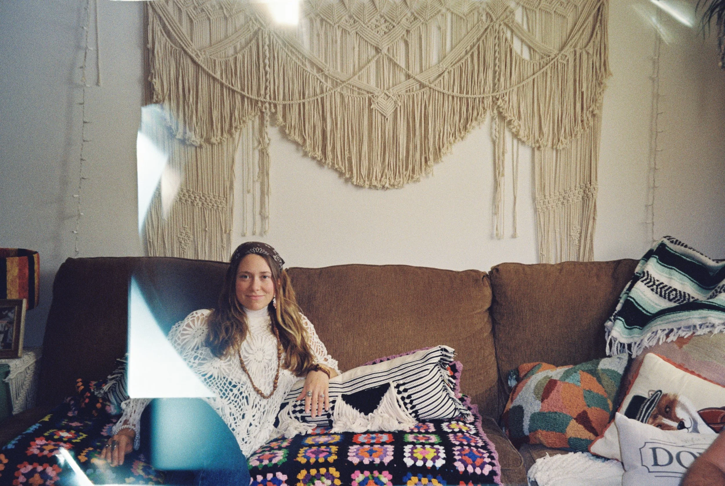 Interior of a room with a brown couch, colorful crochet blanket, patterned pillows, and a large cream-colored macrame wall hanging. A young woman with long wavy hair and a white crochet top is sitting on the couch. Taken by Olivia Ladesic, Wisconsin.