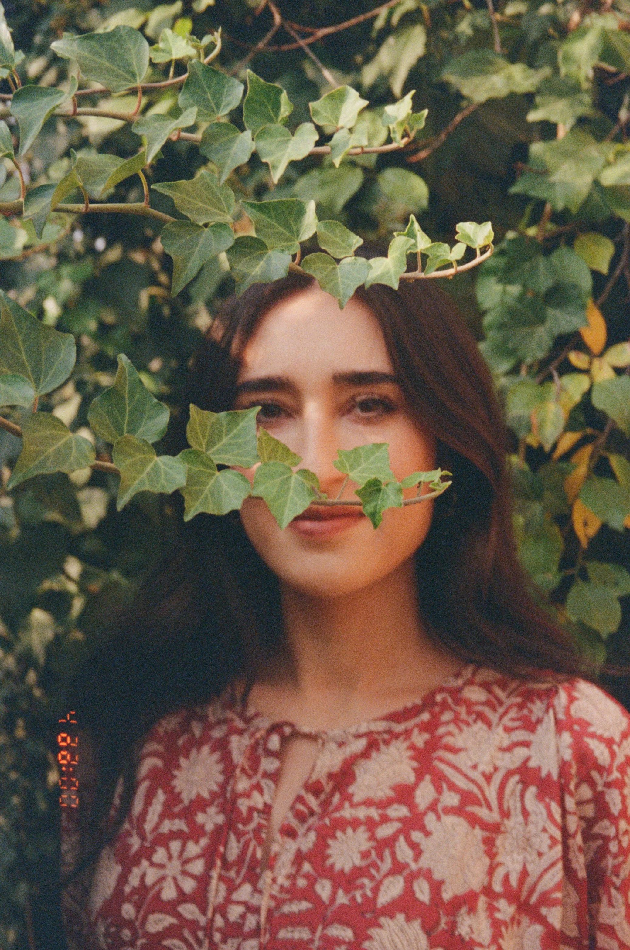 A woman with long, dark hair and a light makeup look, partially visible behind green ivy leaves, with only part of her face showing. Taken by Olivia Ladesic in Tbilisi, Georgia during a portrait photo shoot.