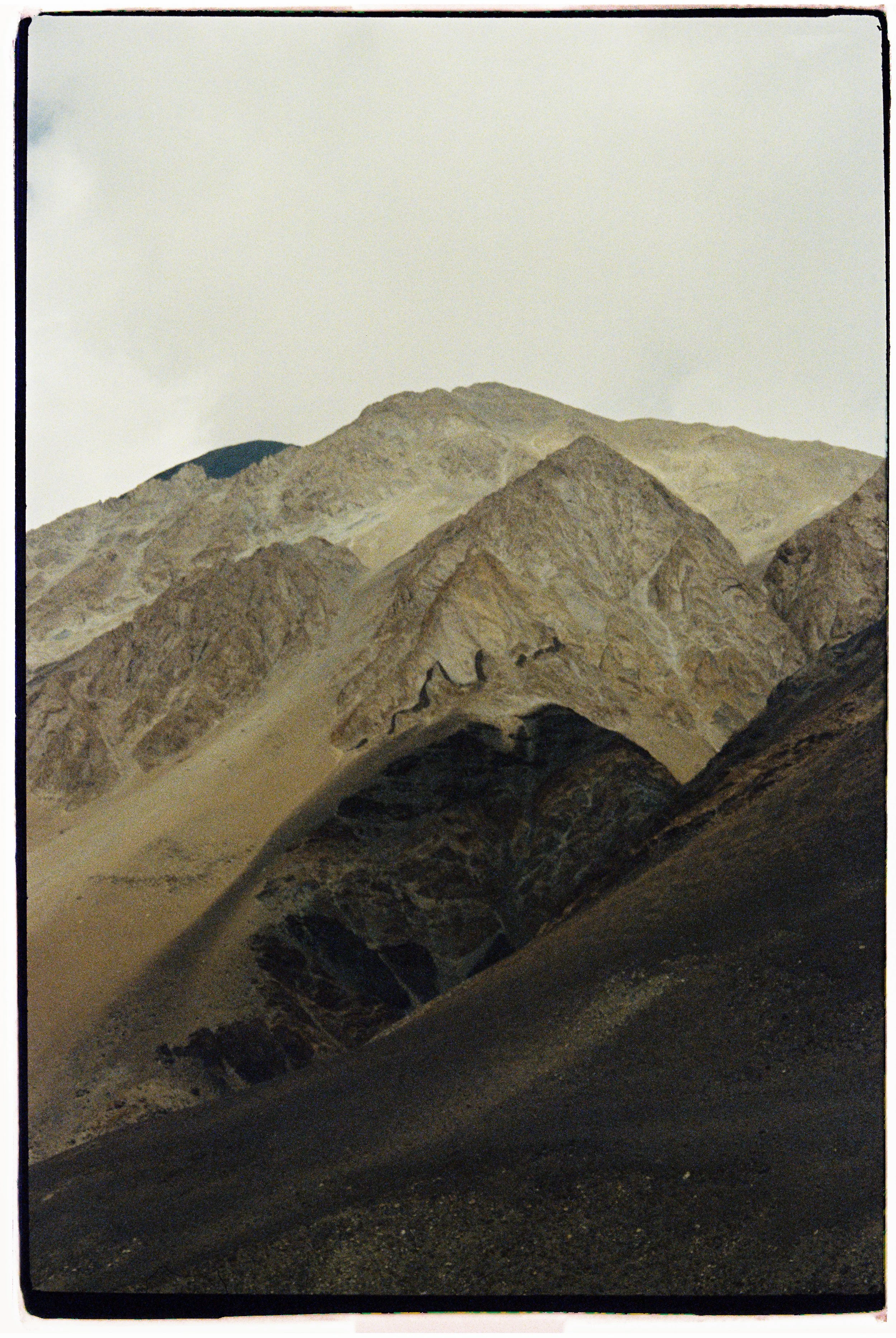 Mountains with rocky, sandy terrain in foreground and peaks in the background under a cloudy sky. Landscape film photography by Olivia Ladesic in Ladakh, India