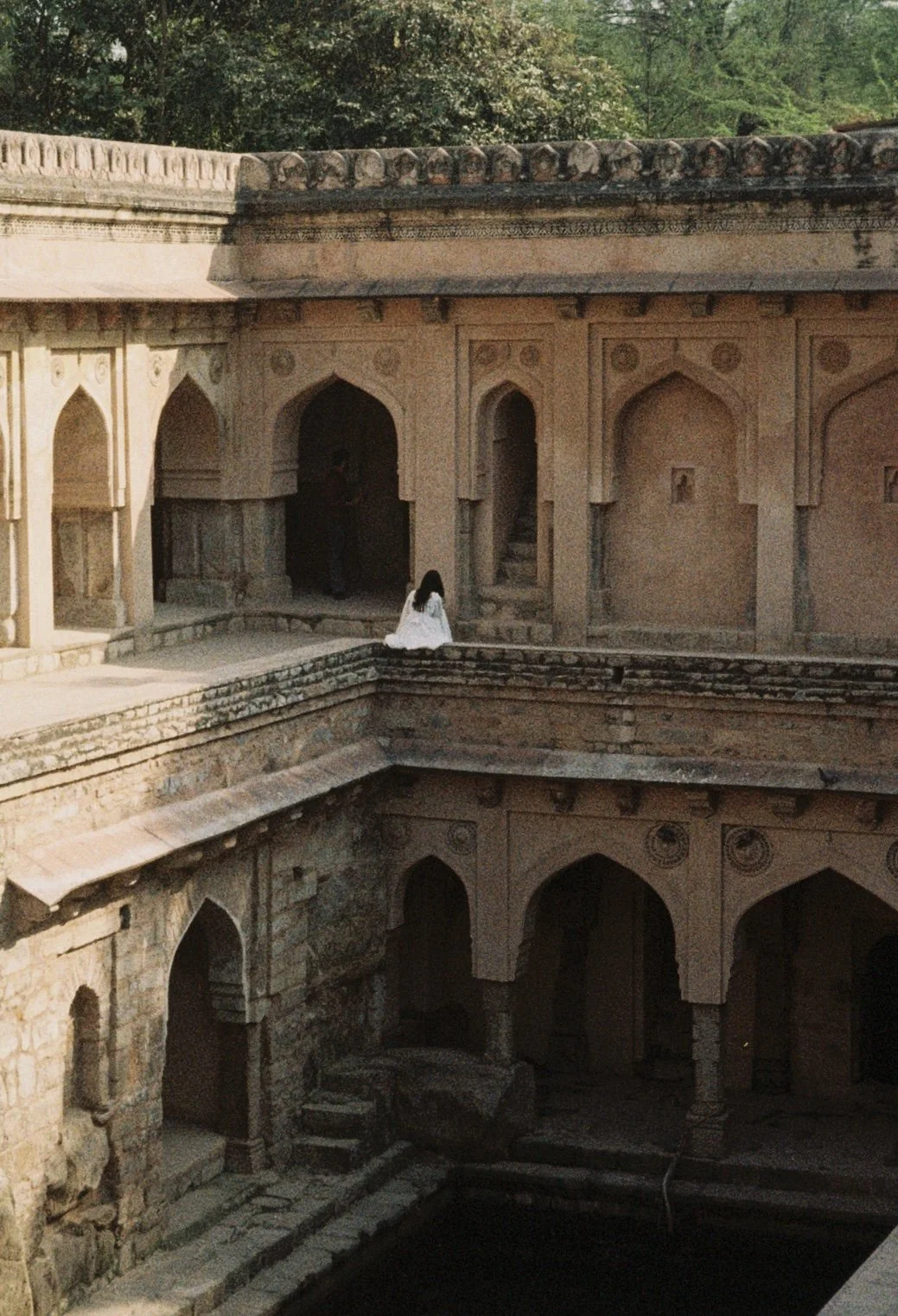 A woman in white dress sitting on the edge of an ancient stone structure with arches, stairs, and intricate carvings, surrounded by greenery. Taken by Olivia Ladesic in New Delhi, India candid film shot.