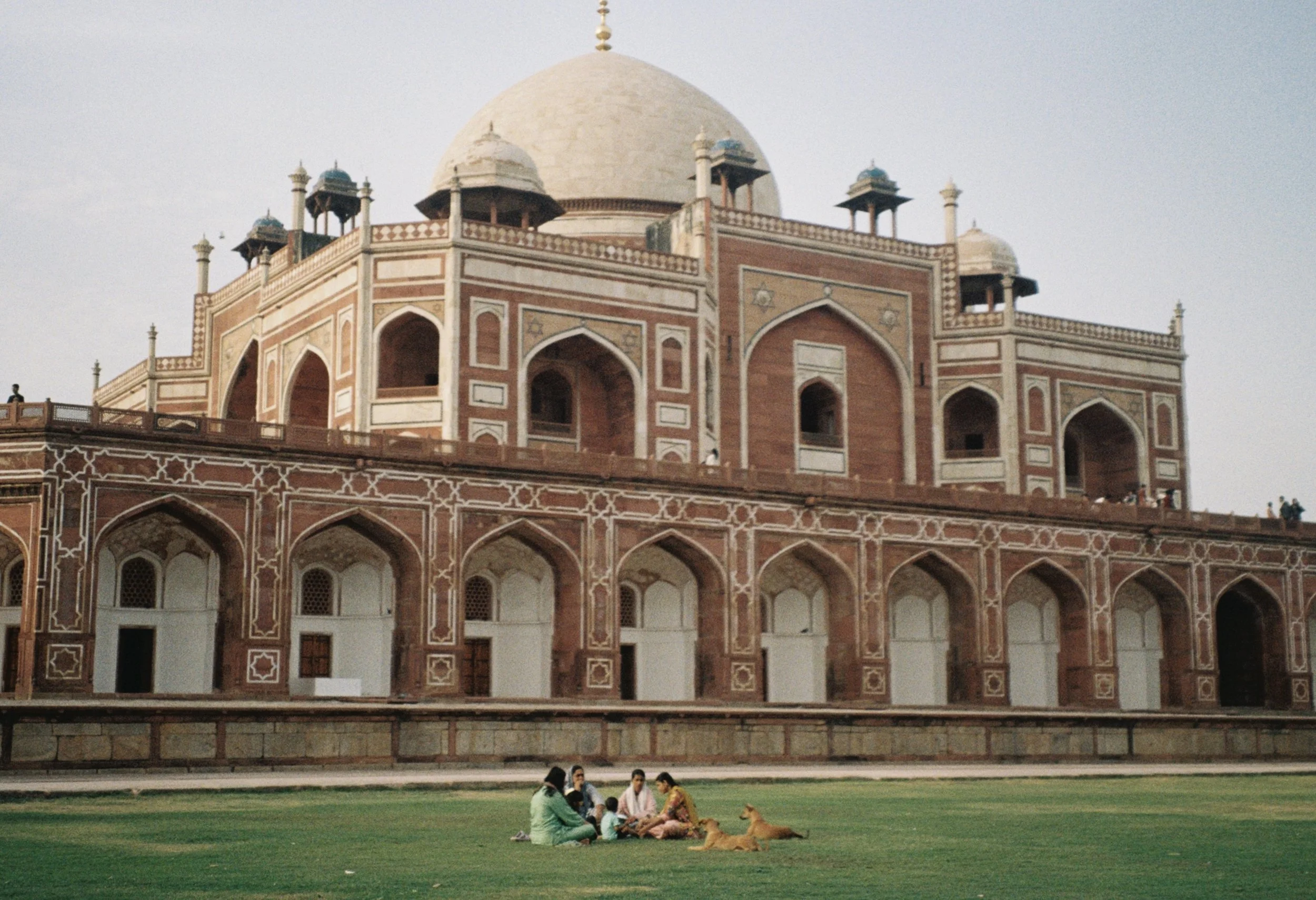 People sitting on green grass in front of the Taj Mahal, India, with a dog, during daylight. Film photo taken by Olivia in New Delhi, India.