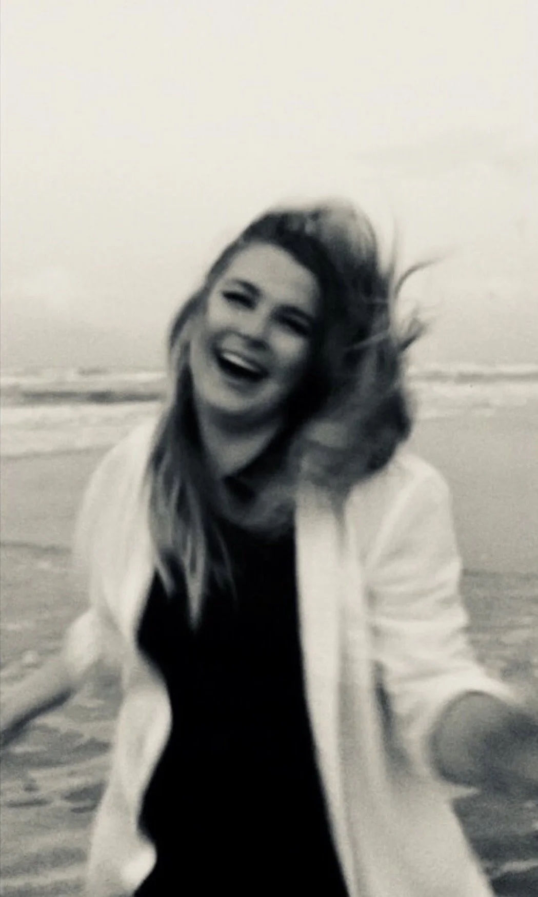 A woman with long hair is smiling and laughing on a beach with the ocean in the background. She is wearing a light-colored jacket over a dark top, and her hair is blowing in the wind. Film photo taken by Olivia Ladesic in Daytona beach, Florida.