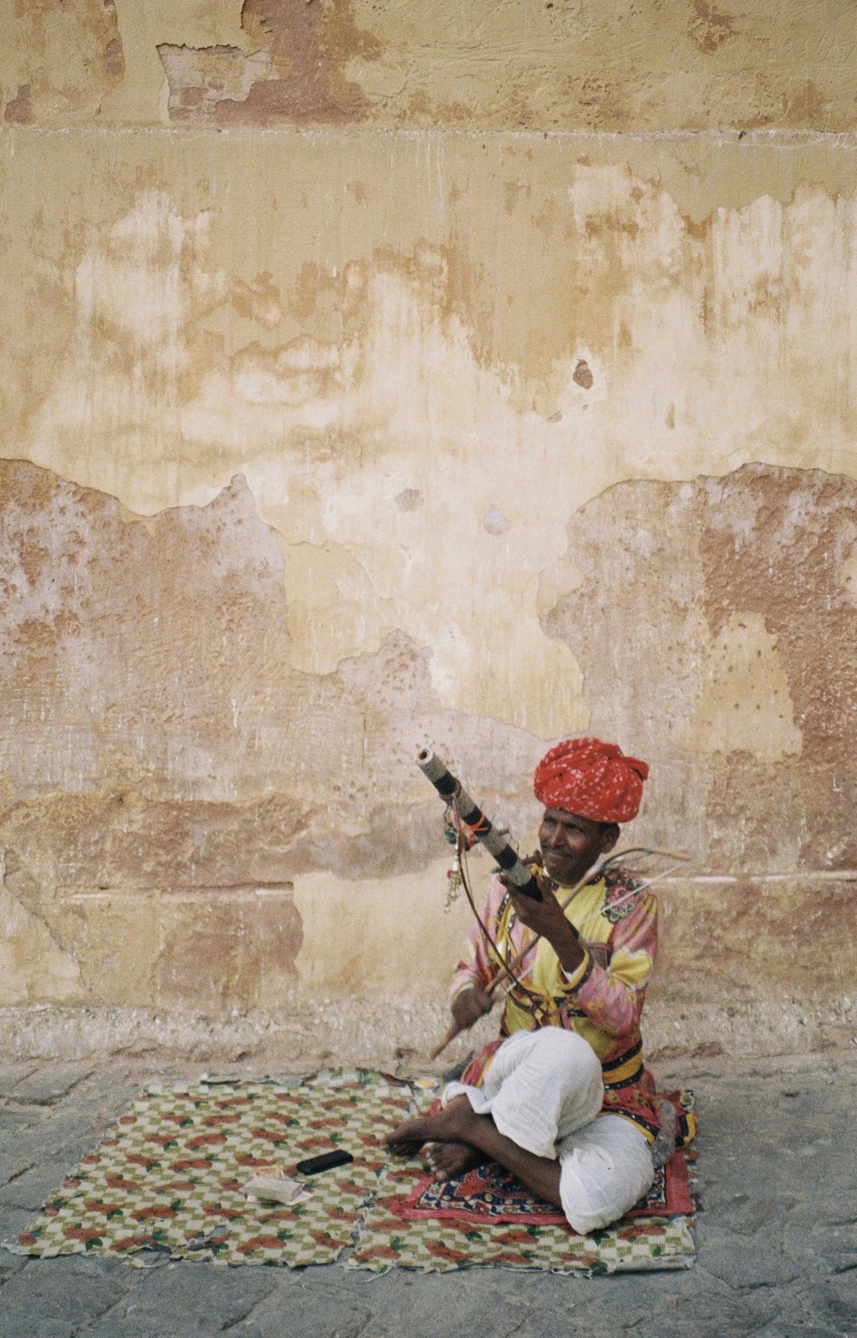 A man sitting on the ground on a patterned mat against a weathered, beige wall. He is wearing a colorful shirt and white pants, holding a long, decorated stick pointed upward, and wearing a red turban. Film photo by Olivia in Jaipur, India