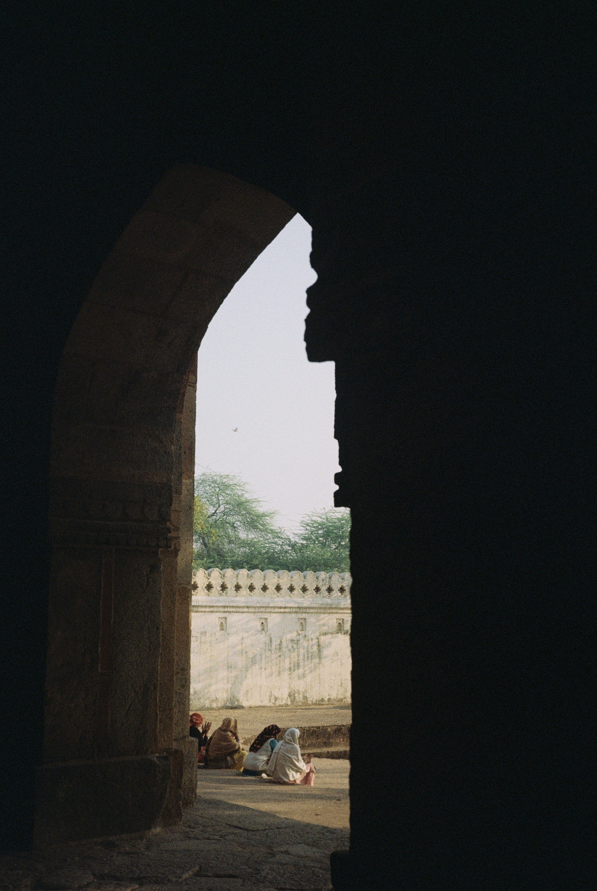 View of a group of people sitting in the courtyard of a historic monument seen through an arched opening with intricate stonework, trees and a white wall in the background. Taken by Olivia Ladesic in Delhi, India on 35mm film.