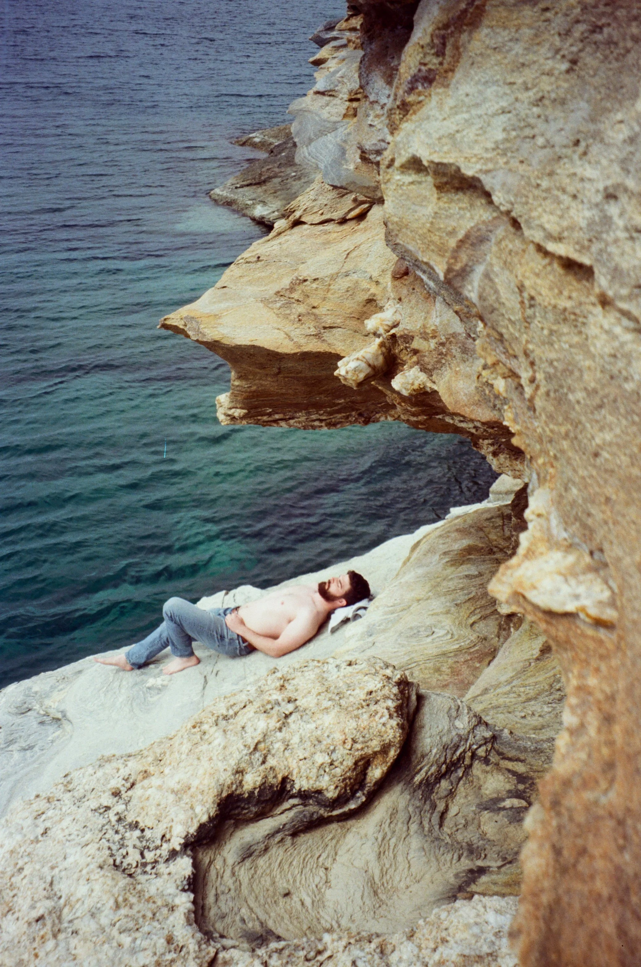 A shirtless man with a beard and jeans lying on a large rock formation by the water, partially shaded by a cliff. Film photo taken by Olivia on a Mediterranean Greek island.