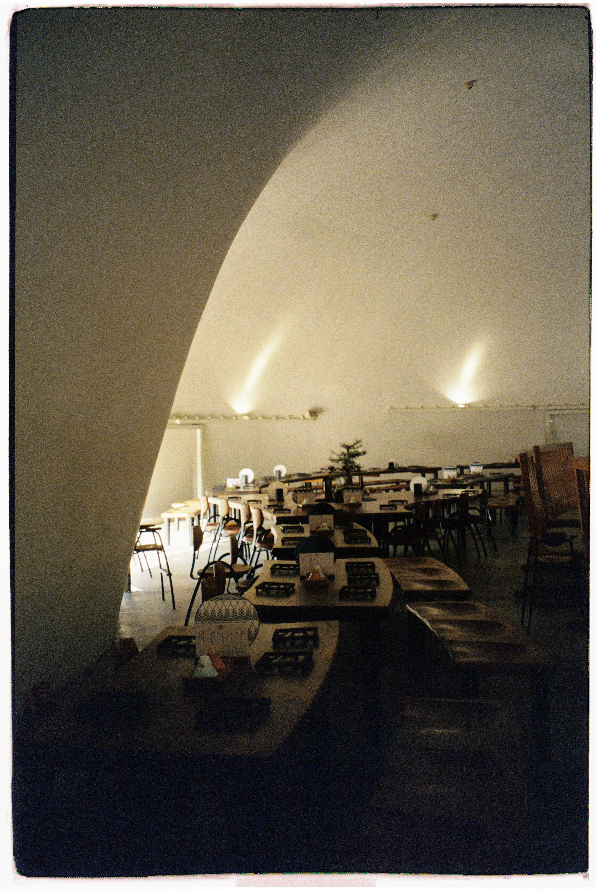 Interior of a restaurant with wooden tables and chairs, some with place settings. Dim lighting and a curved ceiling with artificial light. Film photo taken by Olivia not far from Mt. Fuji, Japan.