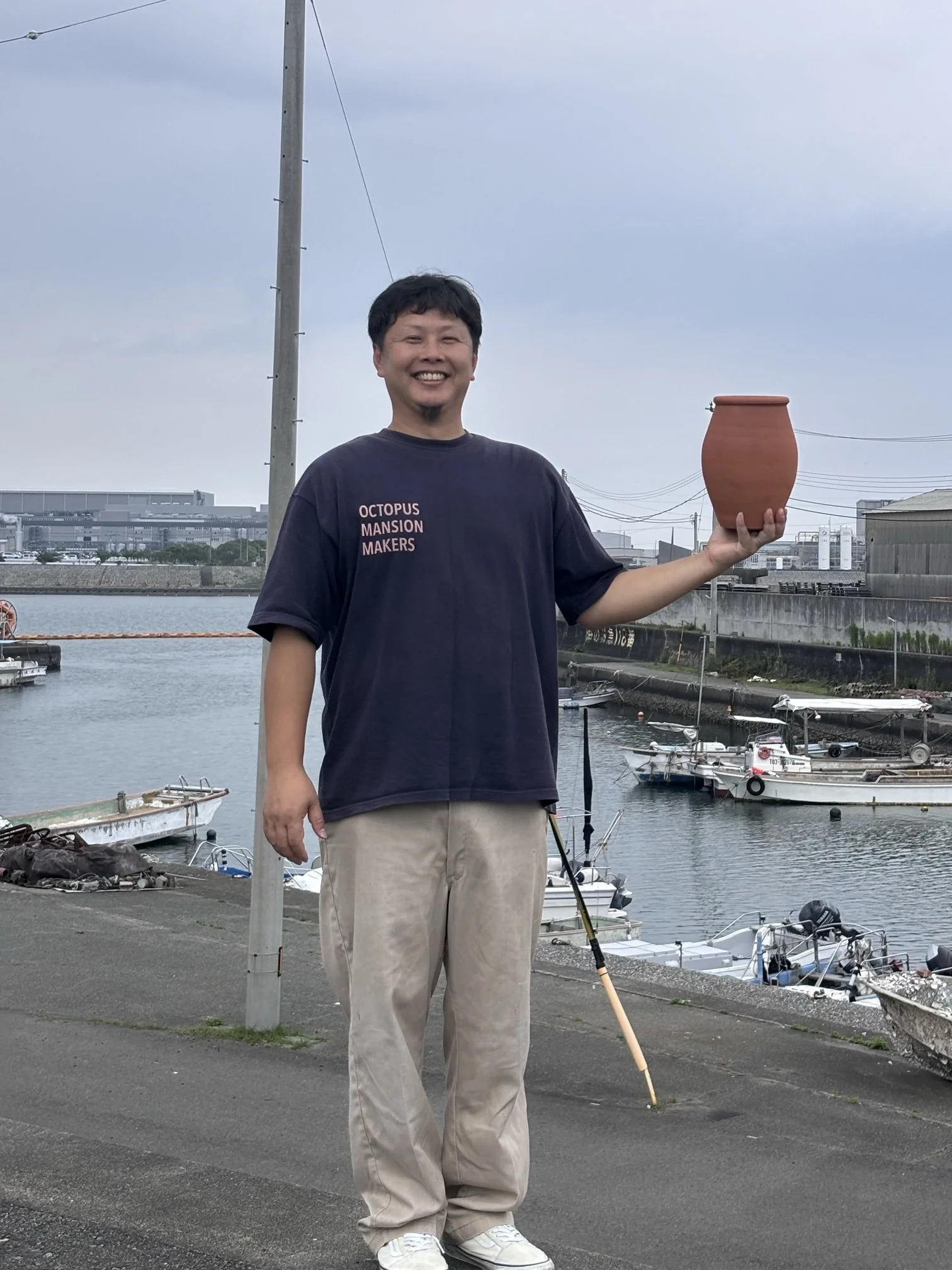 A man standing outdoors near a marina, holding a large clay pot, smiling, with boats and industrial buildings in the background.