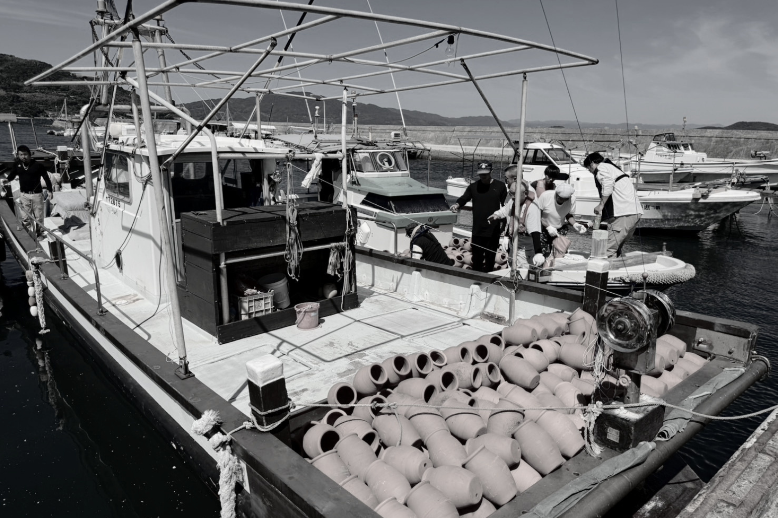 People on a fishing boat handling fishing equipment near other boats docked at a marina.