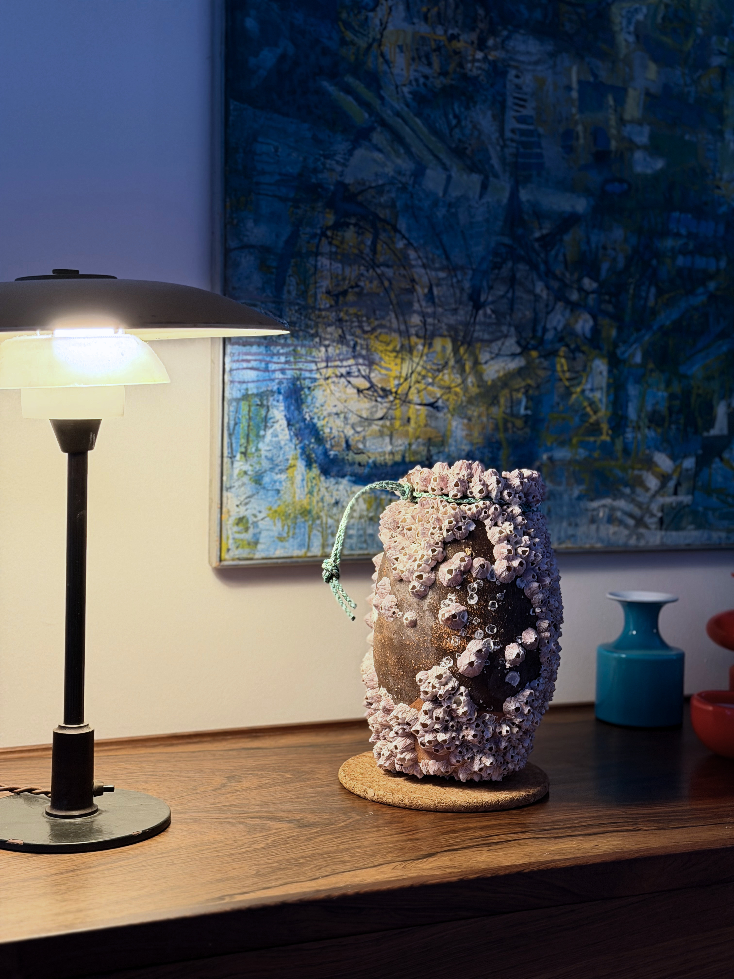 A decorative object made of small shells attached to a rock, mounted on a round base, placed on a wooden table next to a blue vase and a red container, with a painting of a forest scene and a table lamp nearby.