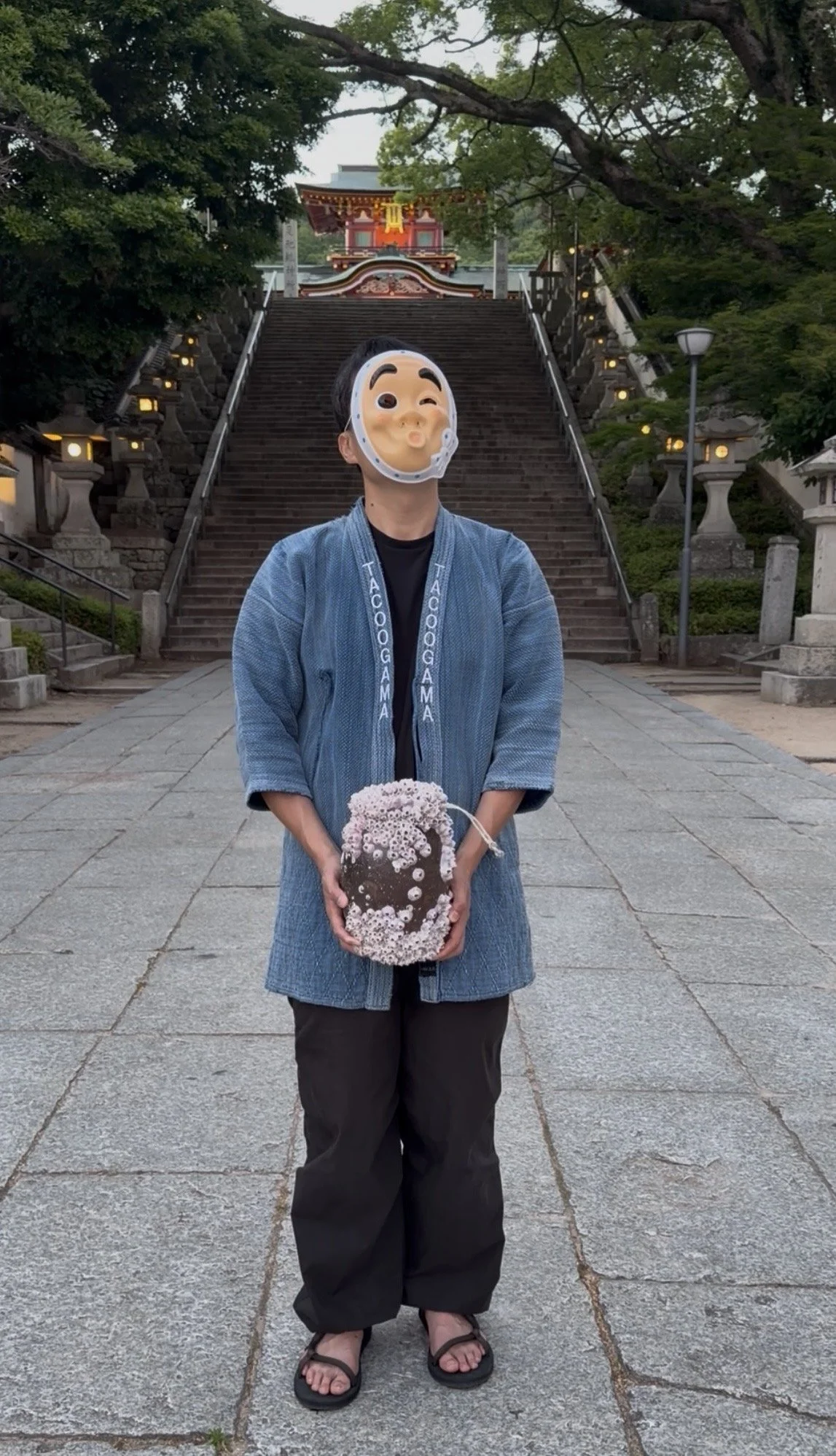 A person wearing a half-face mask and blue traditional Japanese clothing holds a bouquet of flowers in front of a temple with stone steps and lanterns, surrounded by trees.