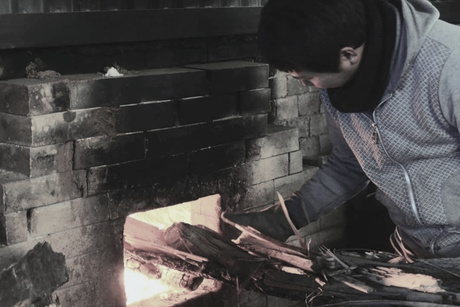 A person kneeling by a brick fireplace, tending to a fire with burning wood.