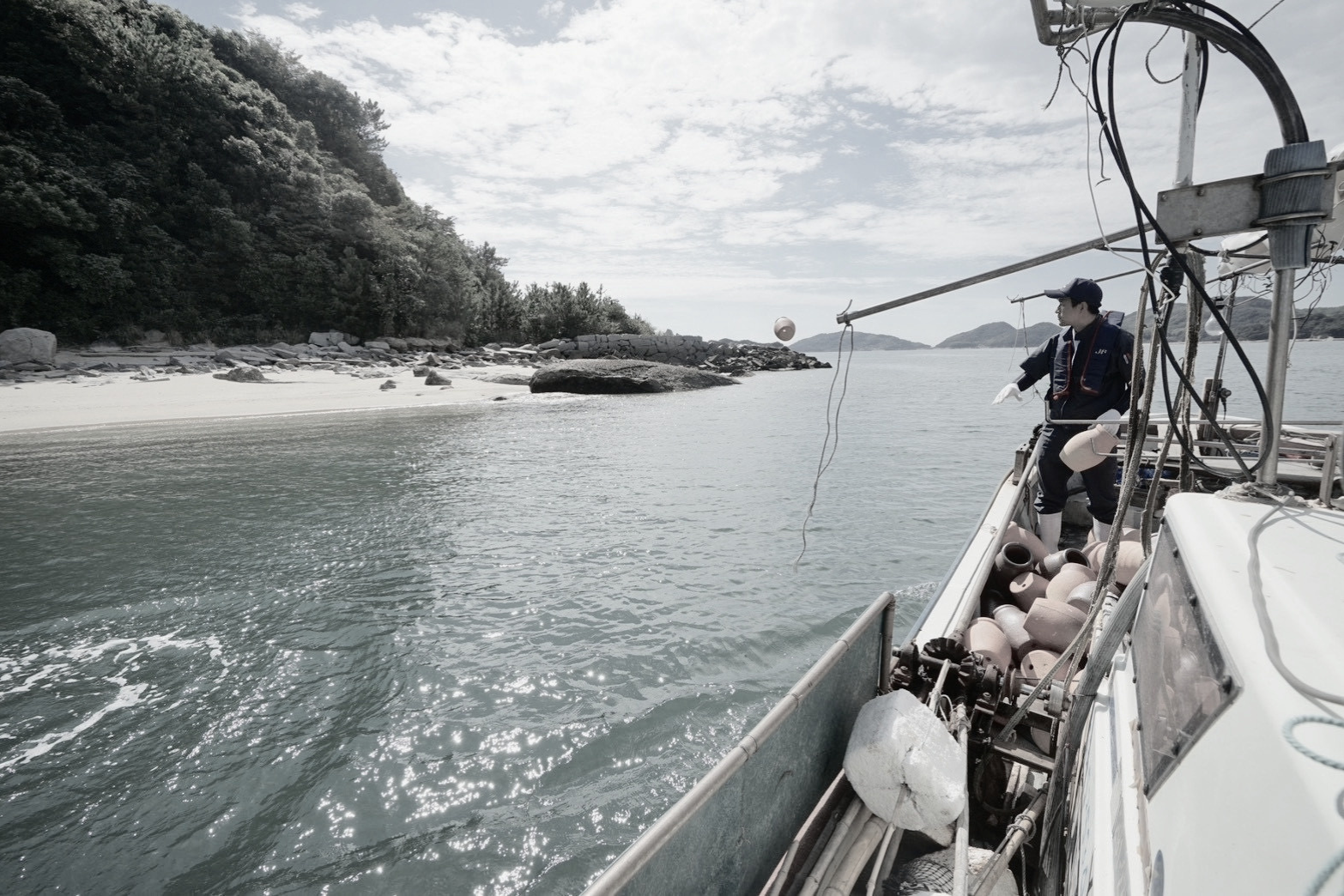 A person on a boat releasing a buoy into the water near a rocky shoreline and wooded hills under a partly cloudy sky.