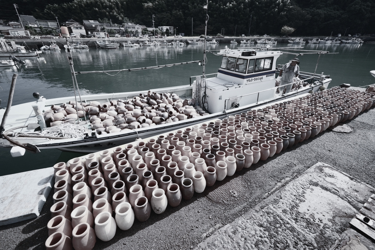 A boat docked at a harbor, filled with numerous traditional Japanese ceramic pots, with more pots lined along the dock, and a scenic background of water, boats, and hillside houses.