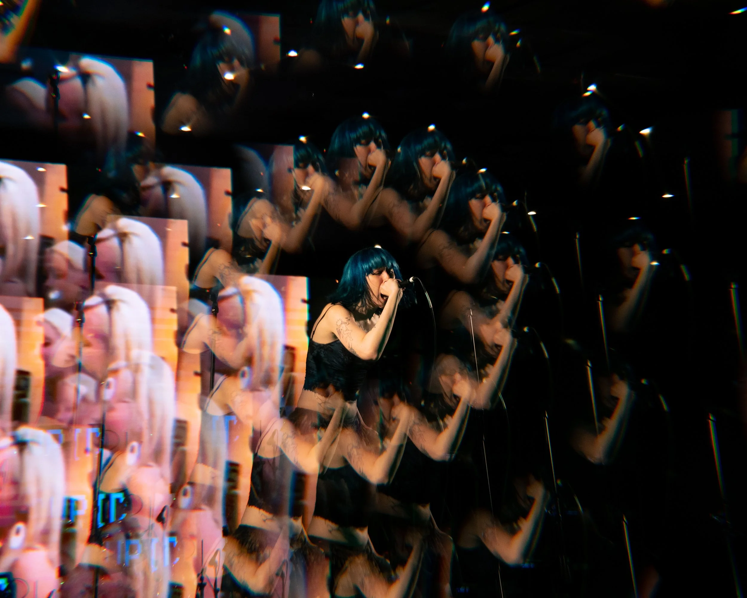 A woman with black hair and tattoos on her arm singing into a microphone on a dark stage, with multiple layered and distorted images of herself creating a glitch effect.