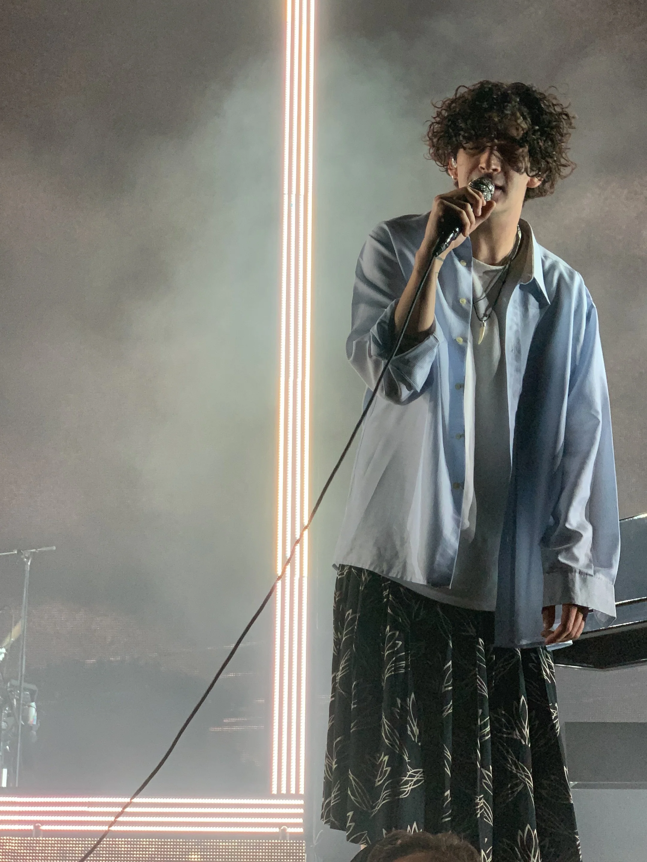 A man with curly hair wearing a blue longsleeve shirt, and a patterned floral skirt, holding a microphone and singing or speaking on a stage with vertical and horizontal light strips in the background.