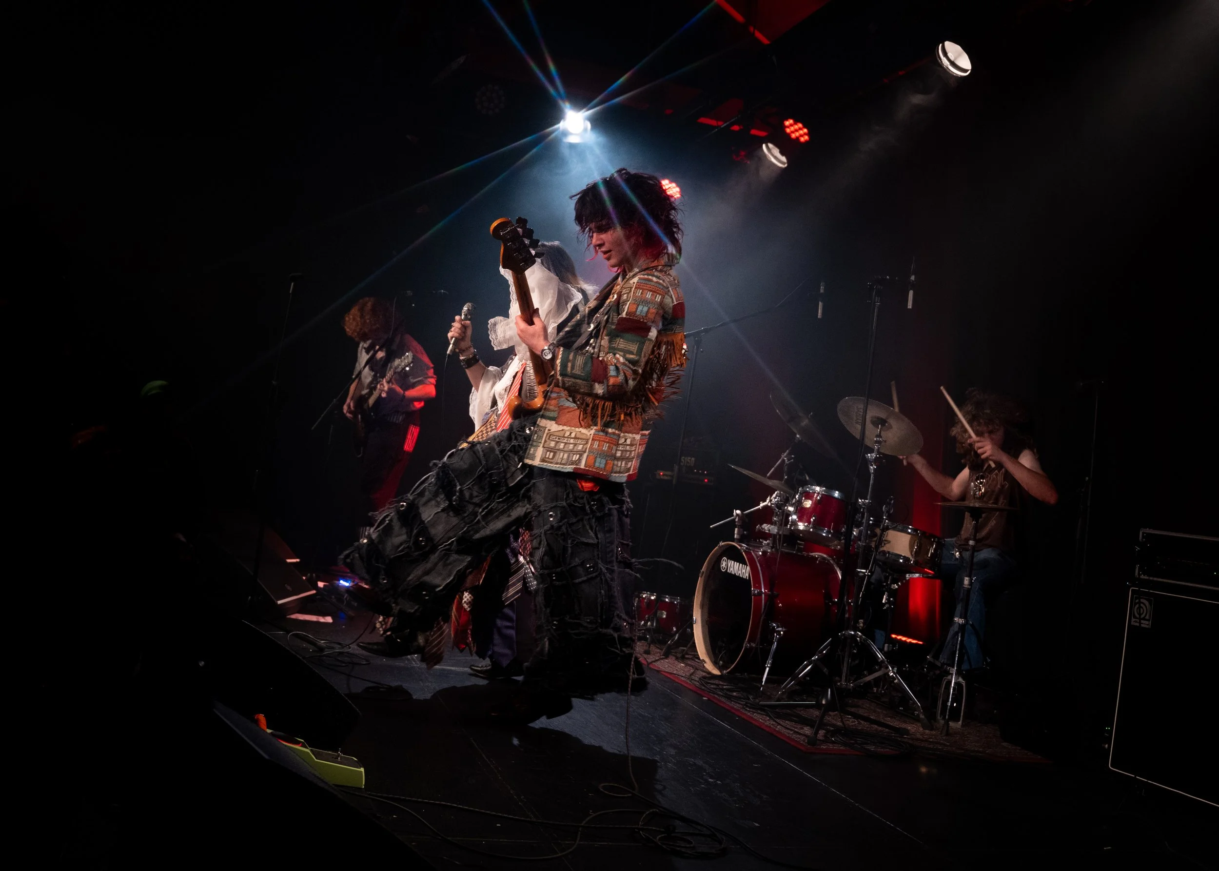 Musicians performing on stage in a dark setting with colorful lights, including a drummer, a guitarist, and a woman singer holding a microphone.