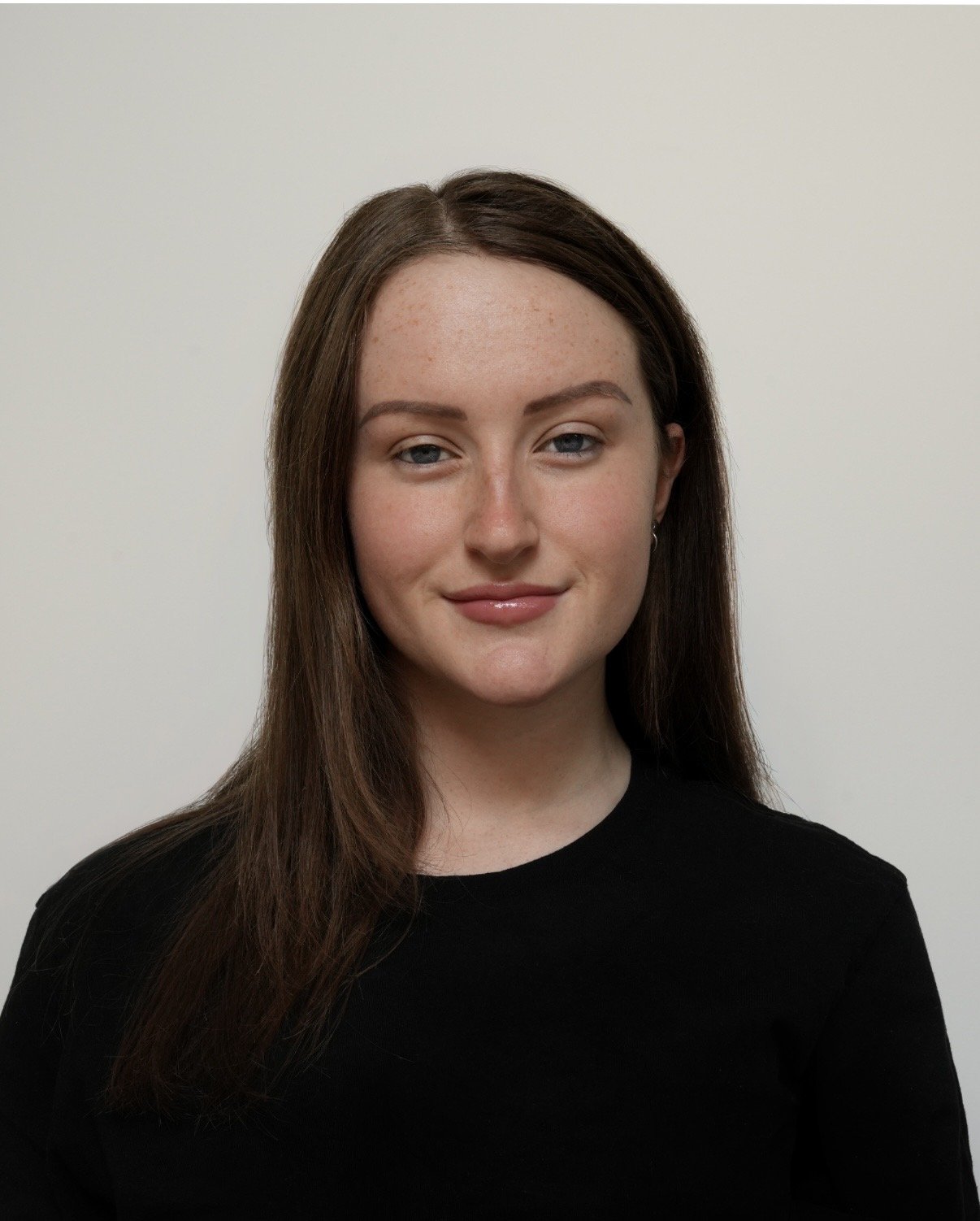 Young woman with long brown hair, wearing a black top, standing against a plain white background.