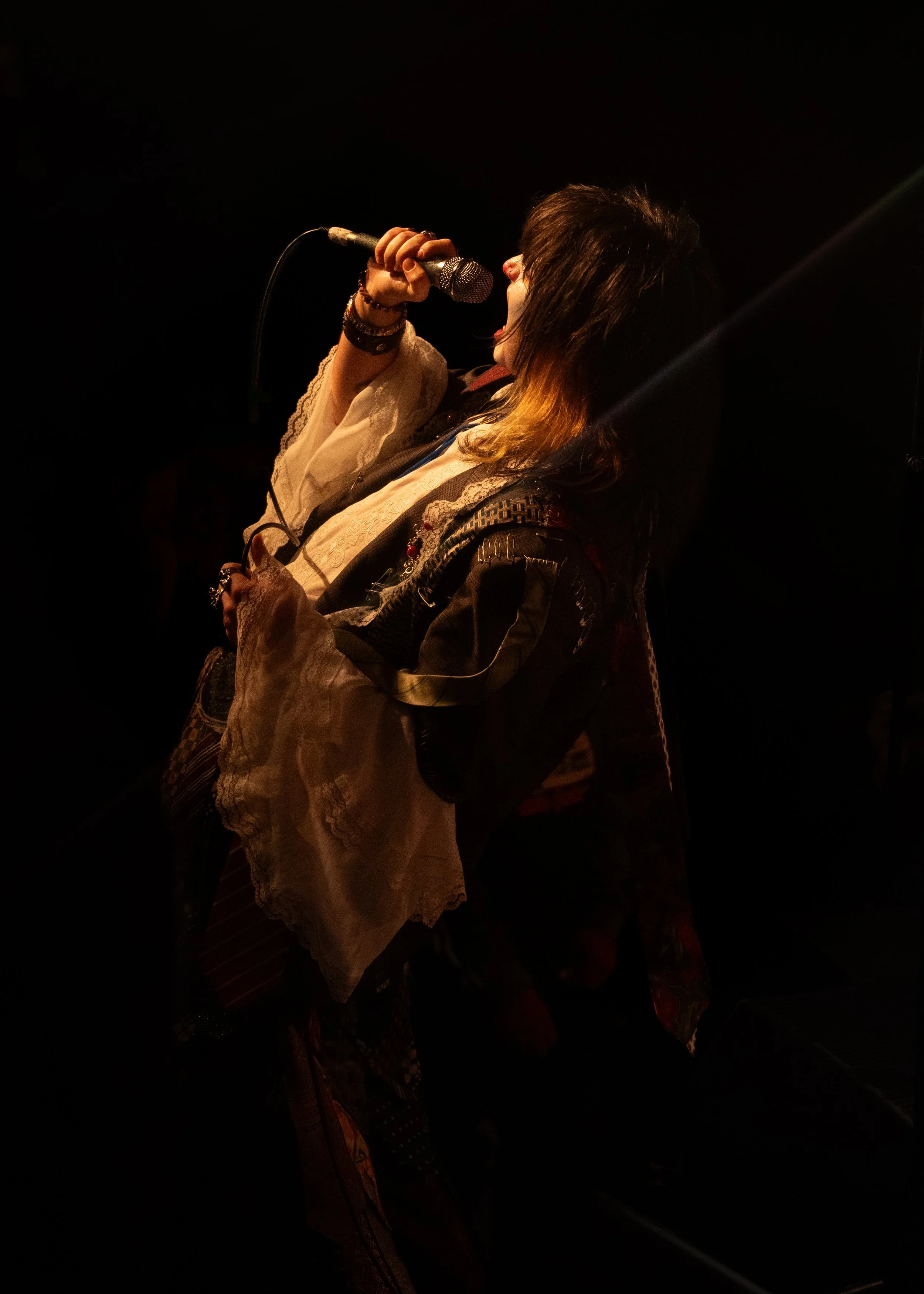 A person singing into a microphone on a dark stage, with light illuminating their face and hands.